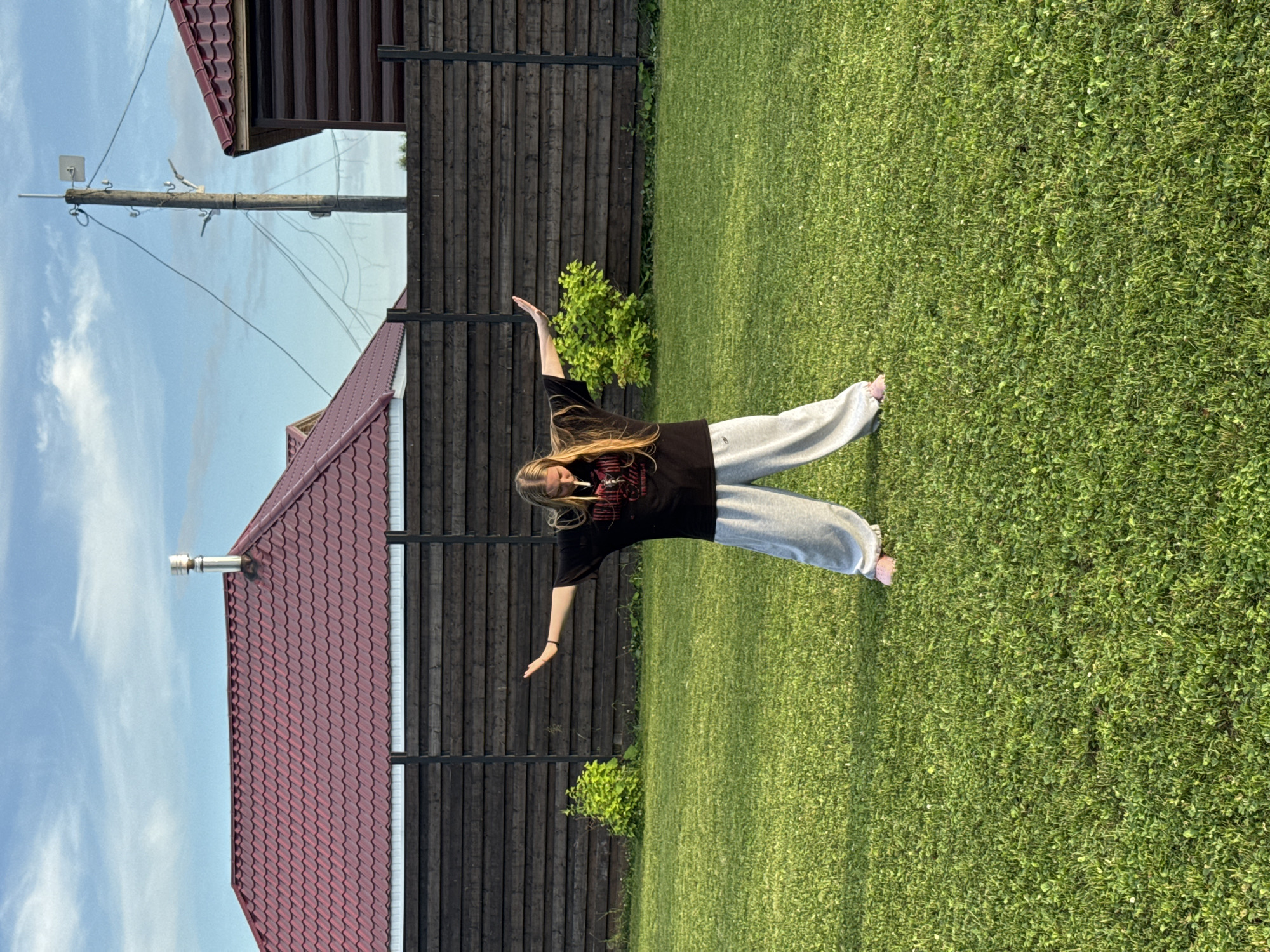 A young woman with blonde hair is standing in a grassy backyard. She is wearing a black t-shirt and loose grey sweatpants. Her arms are outstretched to her sides, and her body is angled slightly to her left. Her head is tilted down, and she appears to be looking at something in her hands. In her mouth, she has what appears to be a cigarette.
The backyard is well-maintained and appears to be in a rural or suburban setting, possibly in the village of Ryabinino, Russia, as suggested by the architecture of the house and fence. Behind the woman, a dark wooden fence runs horizontally across the frame. Beyond the fence, a red-tiled roof of a house is visible, along with a metal chimney. A telephone pole with wires is also present in the background. The sky is blue with some white clouds, and the lighting suggests it is late afternoon or early evening. The grass is green and appears to be a mix of grass and clover.