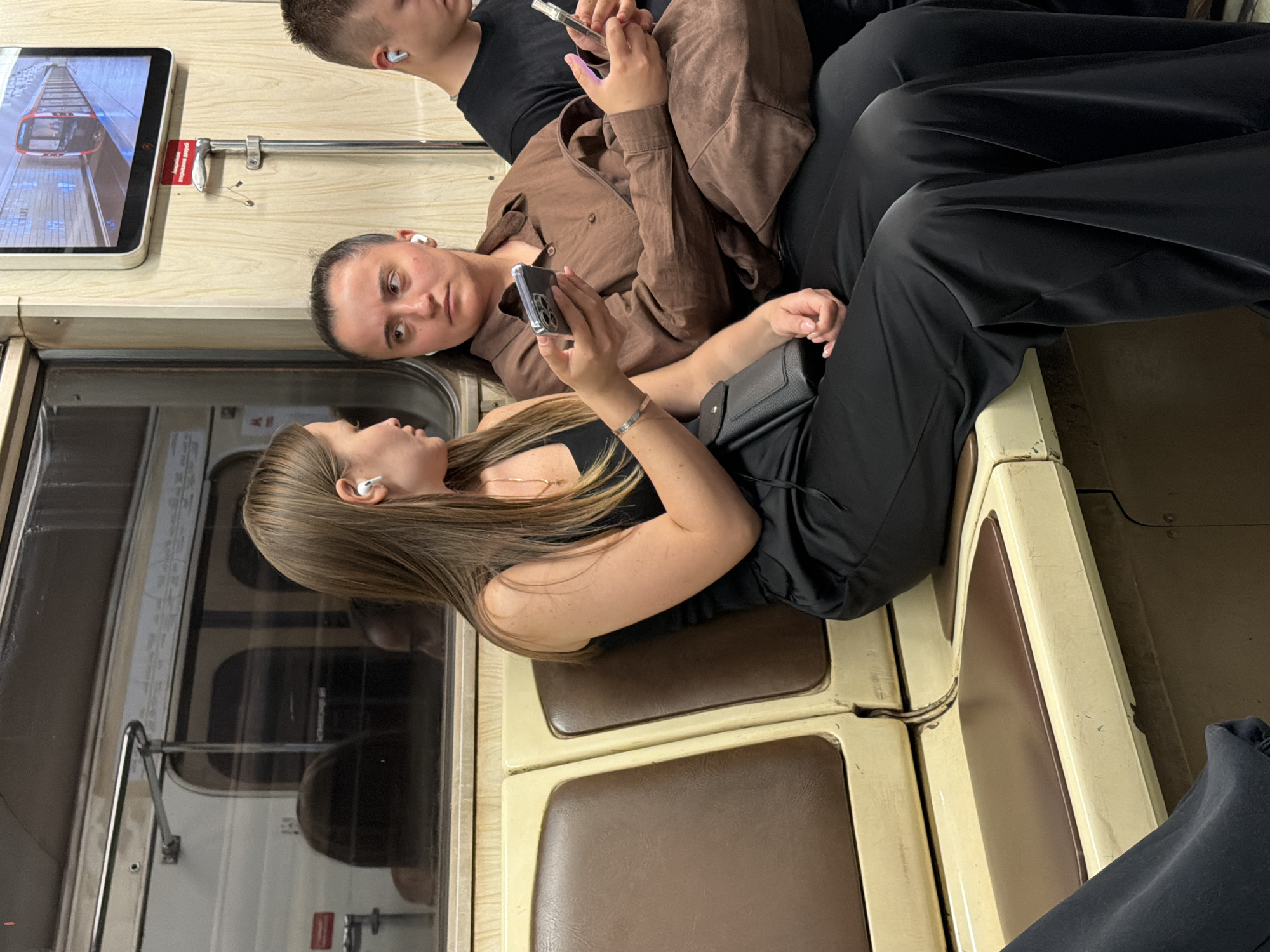 Three individuals are seated in a subway car. On the left, a woman with long brown hair, wearing black attire and white earbuds, holds a smartphone in her right hand. She is looking at the phone. Beside her, a woman with dark hair pulled back, wearing a brown shirt and white earbuds, gazes to her left while holding a smartphone in her right hand. To the right, a person wearing a black t-shirt and white earbuds is partially visible, holding a smartphone. The subway car interior features beige and brown seating and a screen displaying an image of a subway train. The lighting is dim, suggesting an underground environment.