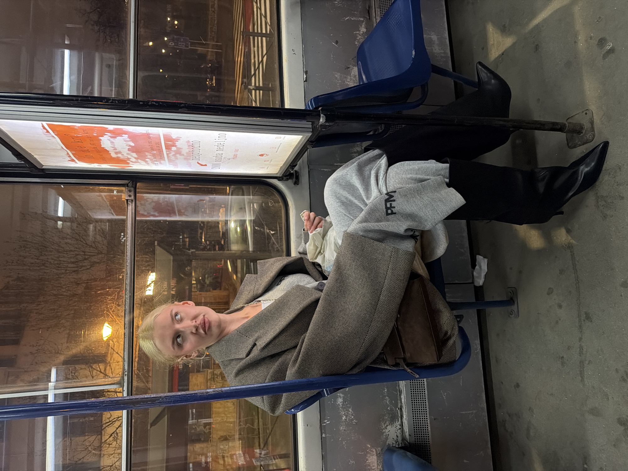 The image depicts the interior of a public transport vehicle, likely a tram, at night in Belgrade, Serbia. A blonde-haired adult female is seated on a blue plastic bucket seat on the left. She wears an oversized brown herringbone patterned blazer over a light grey top, light grey sweatpants with "PFM" printed on the left thigh, and black, high-heeled, pointed-toe boots. A dark brown bag is slung across her body, partially obscured by her blazer. She holds a small, white item in her right hand and looks towards her left (viewer's left) with a somewhat contemplative expression. To her right, an illuminated vertical poster in Serbian advertises blood donation, featuring prominent red text and phrases such as "Ko daje krv?" (Who gives blood?) and "2000 osoba nedeljno" (2000 people weekly), along with a reference to the "Institut za transfuziju krvi Srbije." Through the vehicle's windows, a blurry nighttime cityscape is visible, including streetlights, reflections, and a pedestrian crossing. The vehicle's interior features a grey floor, blue metal support poles, and a second empty blue seat.