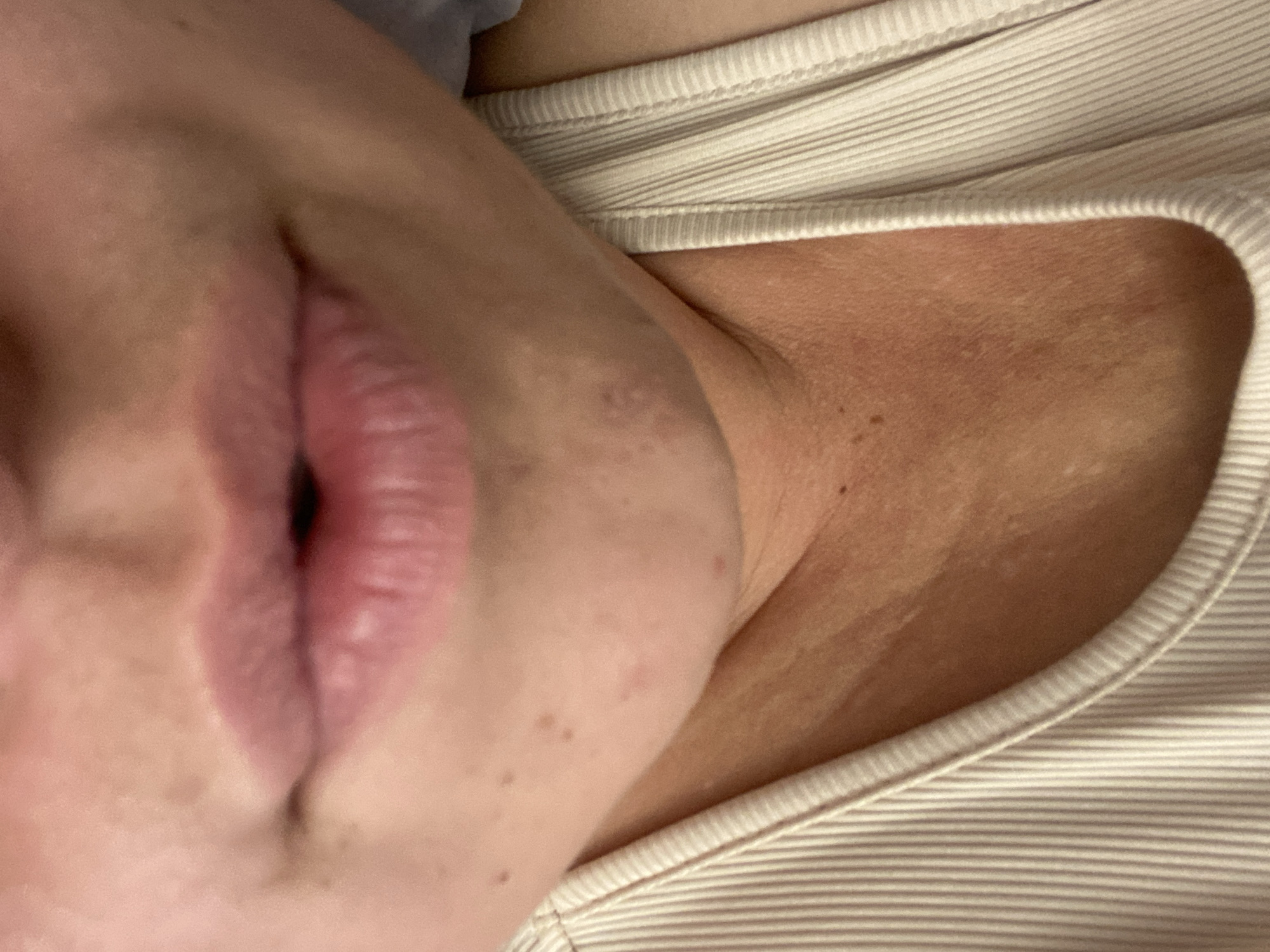 The image is a close-up, low-angle shot of a person's mouth and neck, cropped to show the skin and a portion of a ribbed, cream-colored tank top. The person's lips are slightly parted and appear to be a natural pink color. The skin on the neck shows some texture and faint freckles or moles. The background is out of focus and appears to be a neutral color, possibly a wall or fabric. The lighting is soft, suggesting an indoor setting. There is no visible text, and the time of day, weather, location cues, and emotions are not discernible from the image.