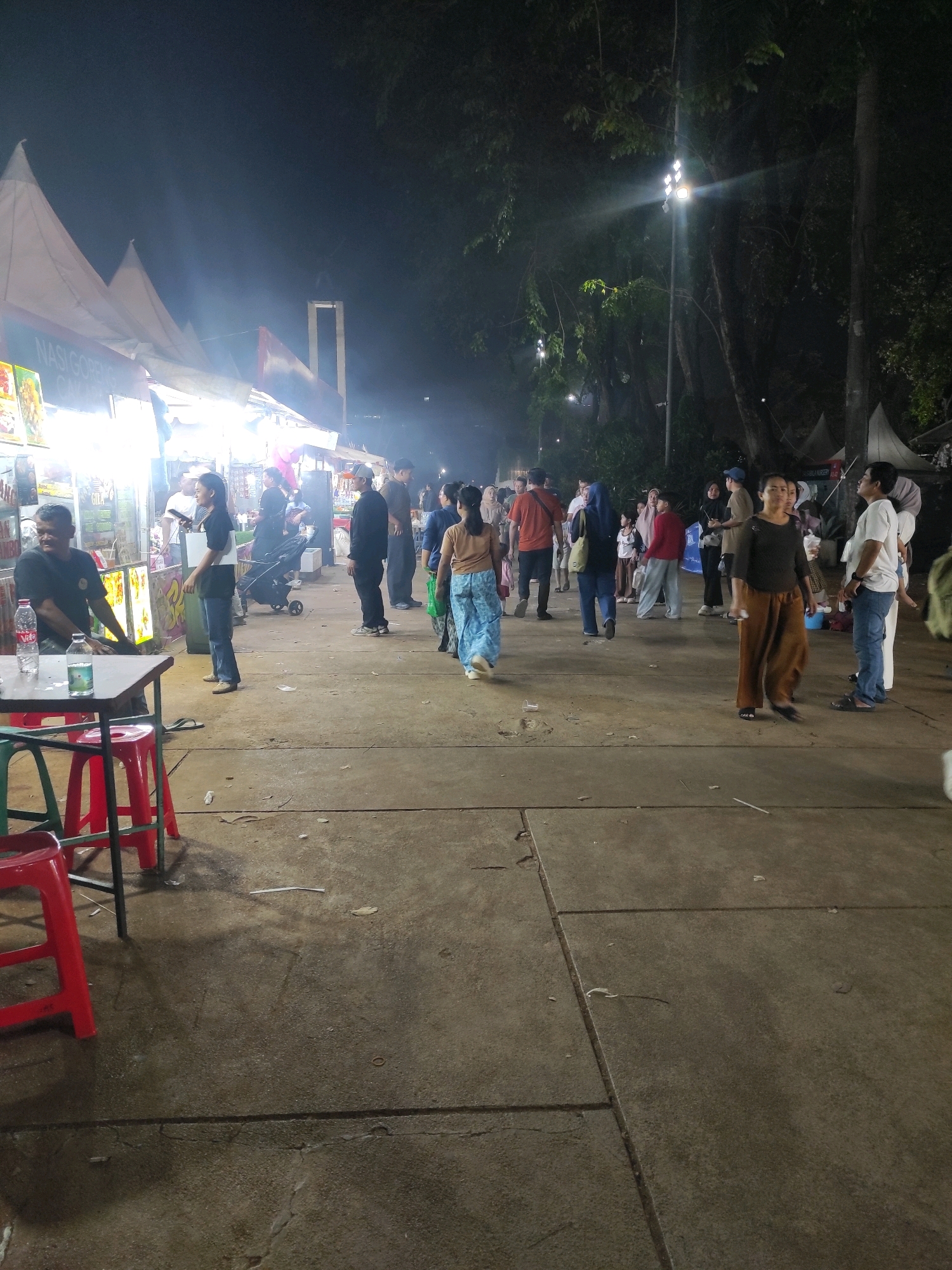 The image depicts an outdoor night scene, likely at a street market or festival, in Central Jakarta, Indonesia. The foreground shows a concrete ground with some debris. A red plastic chair is visible on the left, suggesting a seating area. In the mid-ground, several food stalls with illuminated signs are set up under tents, with smoke emanating from one of them, indicating cooking activity. A man in a black shirt is seated at a table in front of one stall, with bottles of drinks on the table. A woman in a blue shirt and jeans stands near the stall, holding a piece of paper.
A diverse group of people are walking around and interacting in the space between the stalls and the background. They are dressed in casual attire. In the background, trees and streetlights are visible, contributing to the nighttime ambiance. A tall, modern structure resembling a bridge or arch is faintly visible in the distance against the dark sky. The overall atmosphere suggests a lively public gathering.