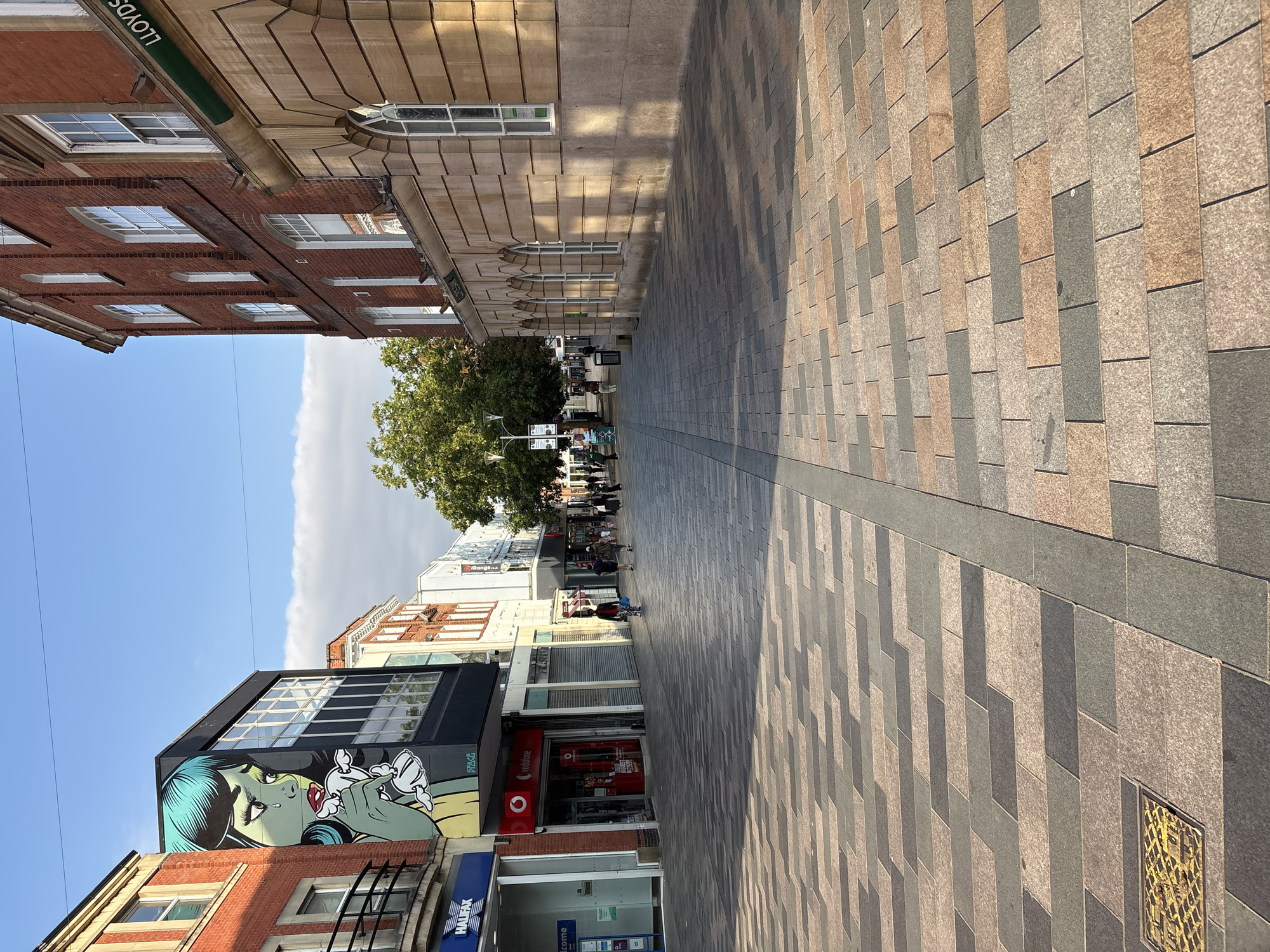 The image depicts a street scene in Chelmsford, United Kingdom. In the foreground, a patterned paved walkway with alternating light brown, dark grey, and greyish-blue rectangular tiles leads into the distance. A metal manhole cover with a yellow grid pattern is visible on the left side of the walkway. The walkway is divided by a darker grey grout line.
On the left side of the street, there are several shopfronts. A prominent feature is a large mural of a crying woman with blue hair on the side of a building. Below the mural, a Halifax bank and a Vodafone store are visible. Further down the street, other shops with closed roller shutters and open entrances are present.
On the right side of the street, a red brick building with stone detailing and arched windows stands. A green sign with "LLOYDS" written on it is affixed to the building. The street is lined with trees, and several individuals are walking along the pavement. The sky above is blue with some scattered clouds, and the sun casts shadows across the scene. The overall impression is that of a commercial street on a clear day.