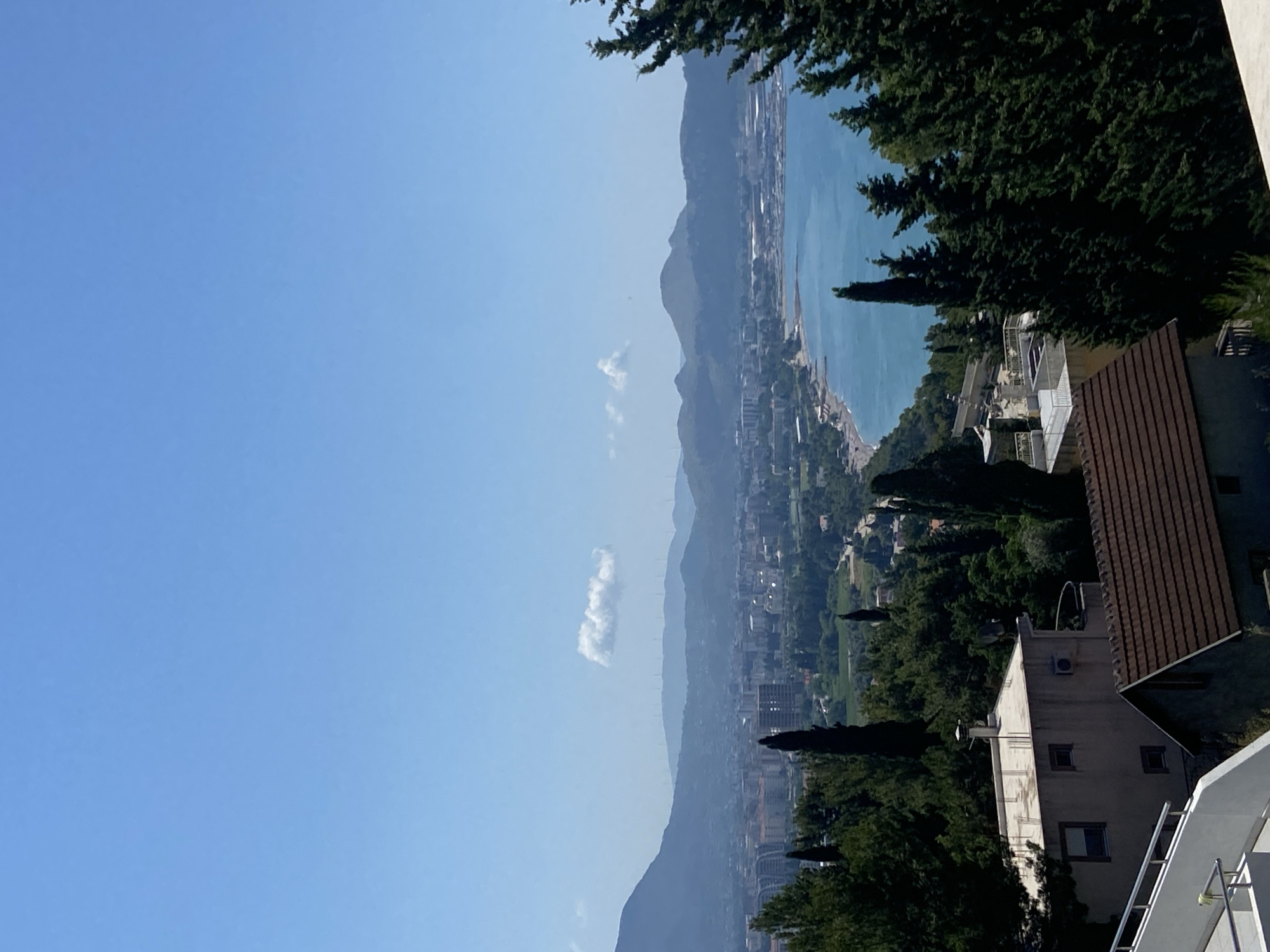 The image captures a scenic view of a coastal city in Šušanj, Montenegro, on a clear, sunny day. The perspective is from a high vantage point, looking down towards the azure sea and a densely populated urban area nestled between rolling hills and mountains. The sky is a brilliant, cloudless blue, indicating fair weather.
In the foreground, the dark green foliage of trees frames the scene, with a portion of a residential building's brown tiled roof and a light-colored house with multiple windows visible. The building appears to have an air conditioning unit mounted on its exterior. Beyond this, the city unfolds with a mix of buildings, some taller and modern, others appearing older. A long stretch of coastline is visible, with a beach or promenade running alongside the sea. The water itself is a vibrant blue, reflecting the clear sky.
Further in the distance, the landscape is dominated by the soft, hazy silhouettes of mountains that rise behind the city. A single, wispy cloud drifts lazily across the sky, adding a touch of depth to the otherwise clear expanse. The overall impression is one of tranquility and natural beauty, with the human settlement harmoniously integrated into the stunning coastal environment. The time of day appears to be late morning or early afternoon, given the bright sunlight and clear visibility. No people or specific activities are discernable, and there is no visible text in the image.