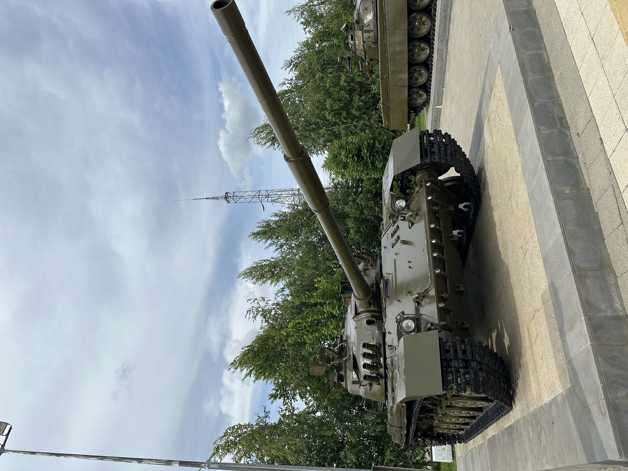 An outdoor, eye-level, medium shot captures two olive-drab tanks on display under a cloudy sky. The tank in the foreground, angled to the left, is the primary subject. Its long cannon extends diagonally upwards and to the right, passing across the frame. The tank exhibits visible tracks, road wheels, headlights, and multiple smaller barrels protruding from the turret. In the background, a second tank, a portion of its side and tracks visible, is positioned to the right. Behind the tanks, a dense line of green trees stretches across the horizon, interspersed with a tall, metallic lattice tower with an antenna at its peak. A lamppost stands on the far left of the frame. The tanks are situated on a light-colored, textured surface bordered by stone-like blocks.