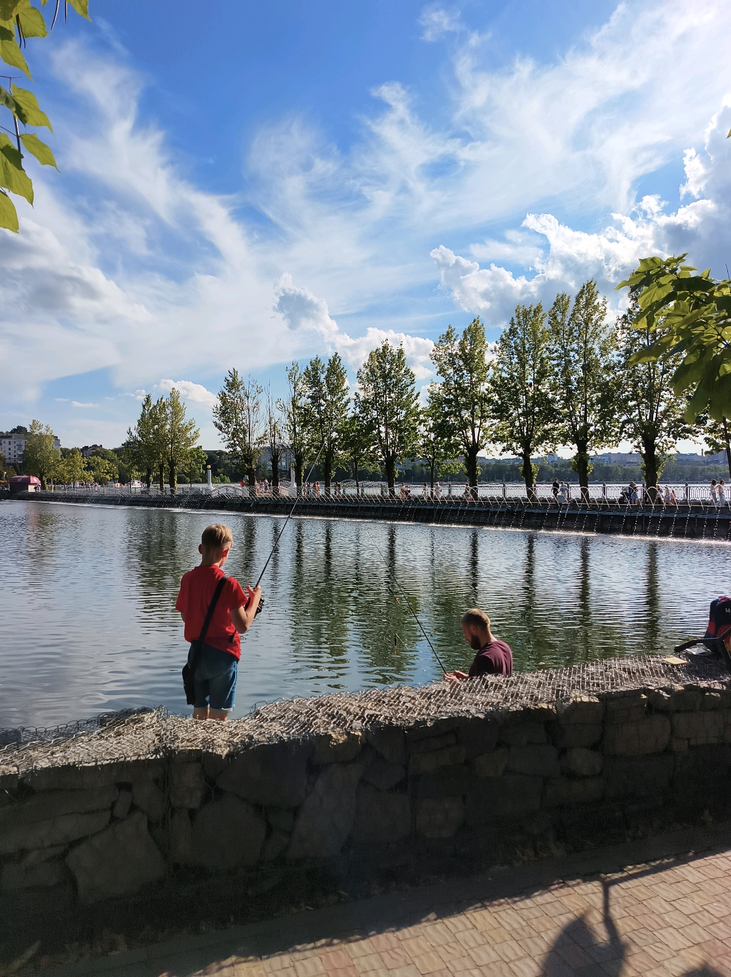 The image depicts a picturesque scene at Ternopil Pond in Ternopil, Ukraine. On the left, a young boy, dressed in a red t-shirt and denim shorts, stands in the water, fishing with a rod. His back is to the camera as he concentrates on his catch. To his right, a man in a maroon t-shirt is also fishing, his focus on his line. Both men are positioned near a gabion wall, a structure made of wire cages filled with stones, which lines the edge of the pond.
The pond itself is calm, with reflections of the trees and the sky shimmering on its surface. Across the pond, a tree-lined embankment is visible, with a promenade where several people are walking and enjoying the day. Beyond the promenade, a city skyline with buildings and structures suggests an urban environment. The sky above is a vibrant blue, dotted with fluffy white clouds, some of which are wispy and stretched, characteristic of fair weather. The overall atmosphere is serene, capturing a moment of leisure and connection with nature within the city.