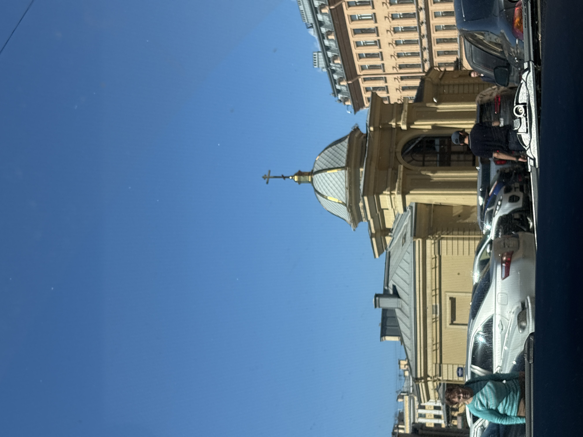 The image depicts a street scene in Saint Petersburg, Russia. In the foreground, a dark blue car windshield is visible, with a reflection on it. Behind the windshield, several cars are parked on the street, including a white sedan and a black SUV. A woman wearing a turquoise striped sweater and brown pants stands to the left of the white sedan. To the right of the black SUV, a man wearing a dark blue t-shirt and a grey baseball cap is seen from the back. Further back, a building with a golden dome topped with a cross is visible. This building appears to be an Orthodox church, potentially the Church of the Resurrection of Christ or a similar religious building given its architectural style and the presence of a cross. To the right of the church is a multi-story building with numerous windows and a light orange facade. The sky is clear and blue. The overall environment suggests an urban setting in Saint Petersburg, Russia, on a sunny day.