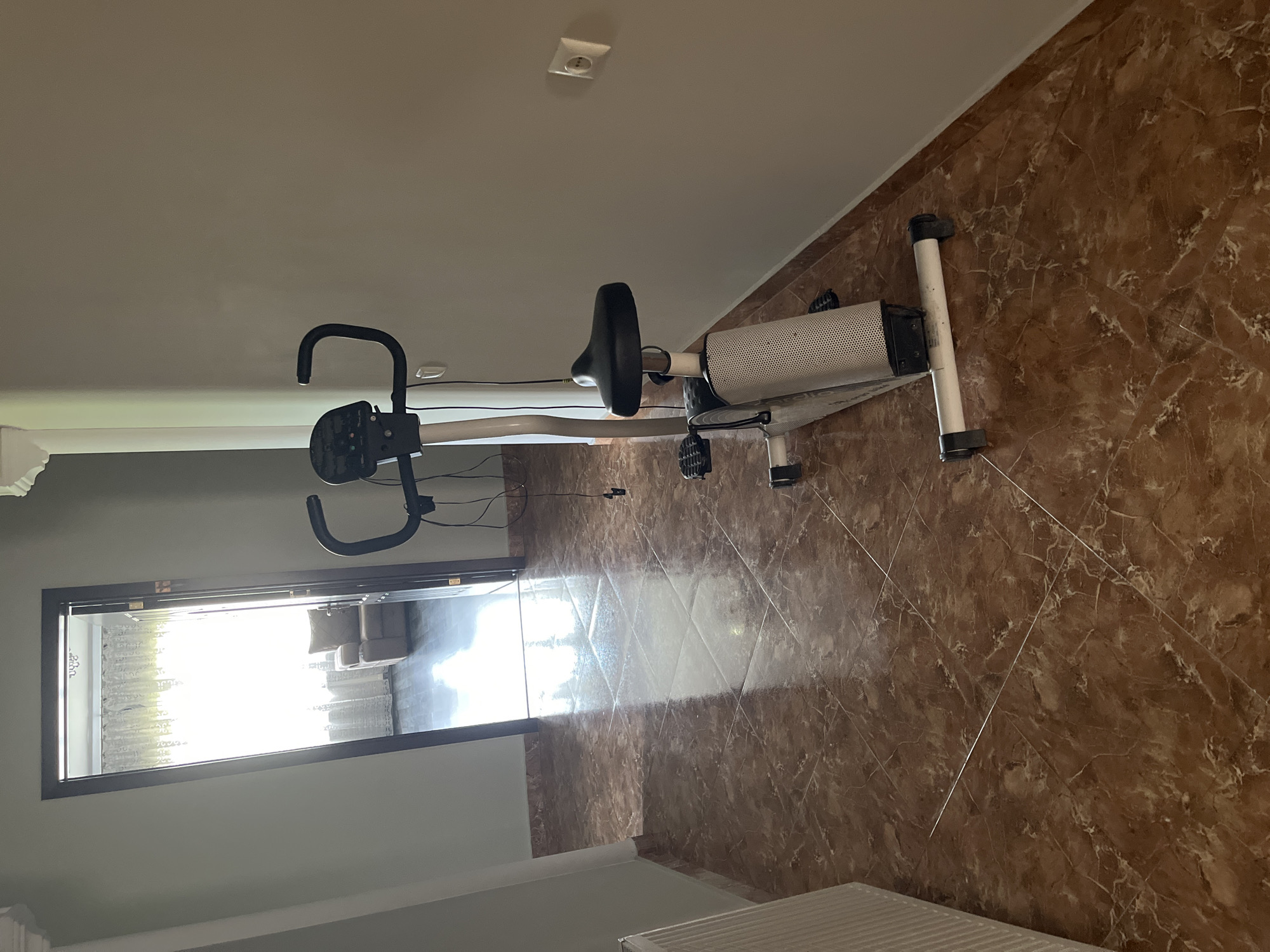 An indoor, eye-level, full shot captures an exercise bike positioned in a hallway. The bike is white and black with a digital display and black handlebars. The hallway features marble flooring and light gray walls. To the left, a white radiator is visible. An open doorway on the left reveals a glimpse of another room with a single armchair and a large window covered by white curtains. An electrical outlet is present on the right wall. The lighting suggests daylight.