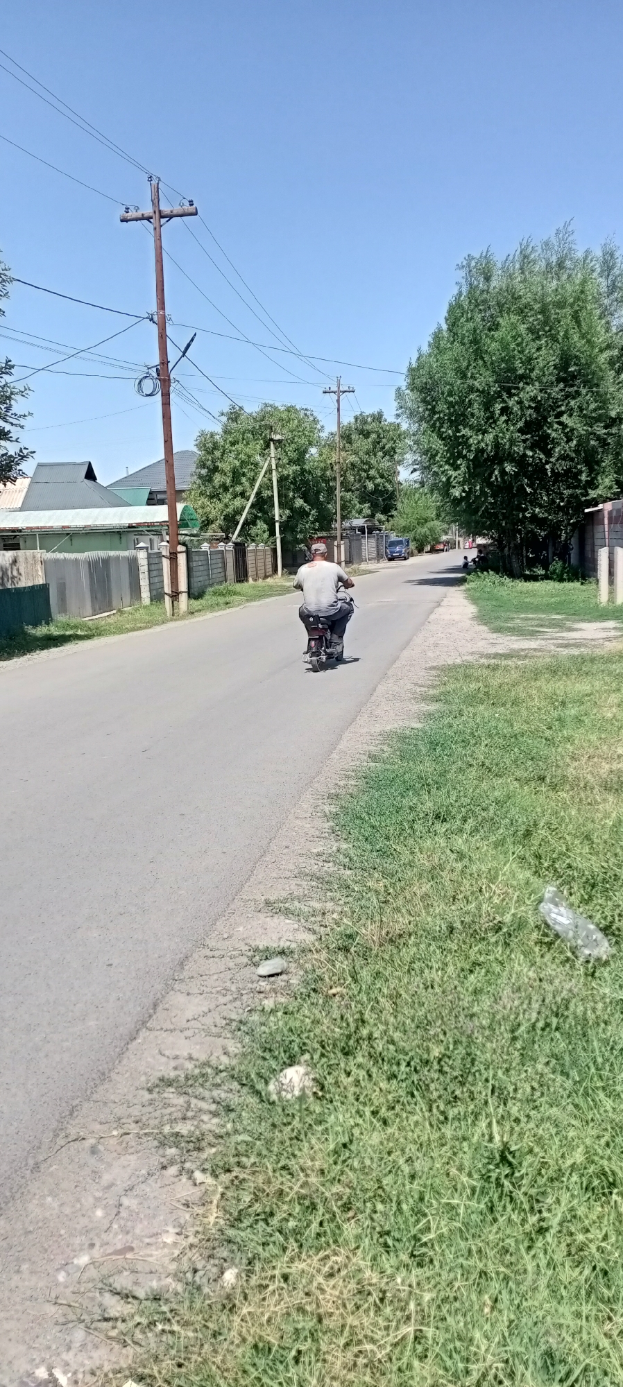 In Sokuluk, Kyrgyzstan, a man rides a motorcycle on a paved road. The man, wearing a light-colored shirt and a cap, is positioned in the center of the road, moving away from the viewer. The road is flanked by grassy verges, one of which shows some scattered debris, including a plastic bottle. On the left side of the road, there are fences and houses with pitched roofs, characteristic of a residential area. Overhead, power lines are suspended between wooden utility poles. The sky is clear and blue, indicating a sunny day. The environment suggests a rural or suburban setting within the city.