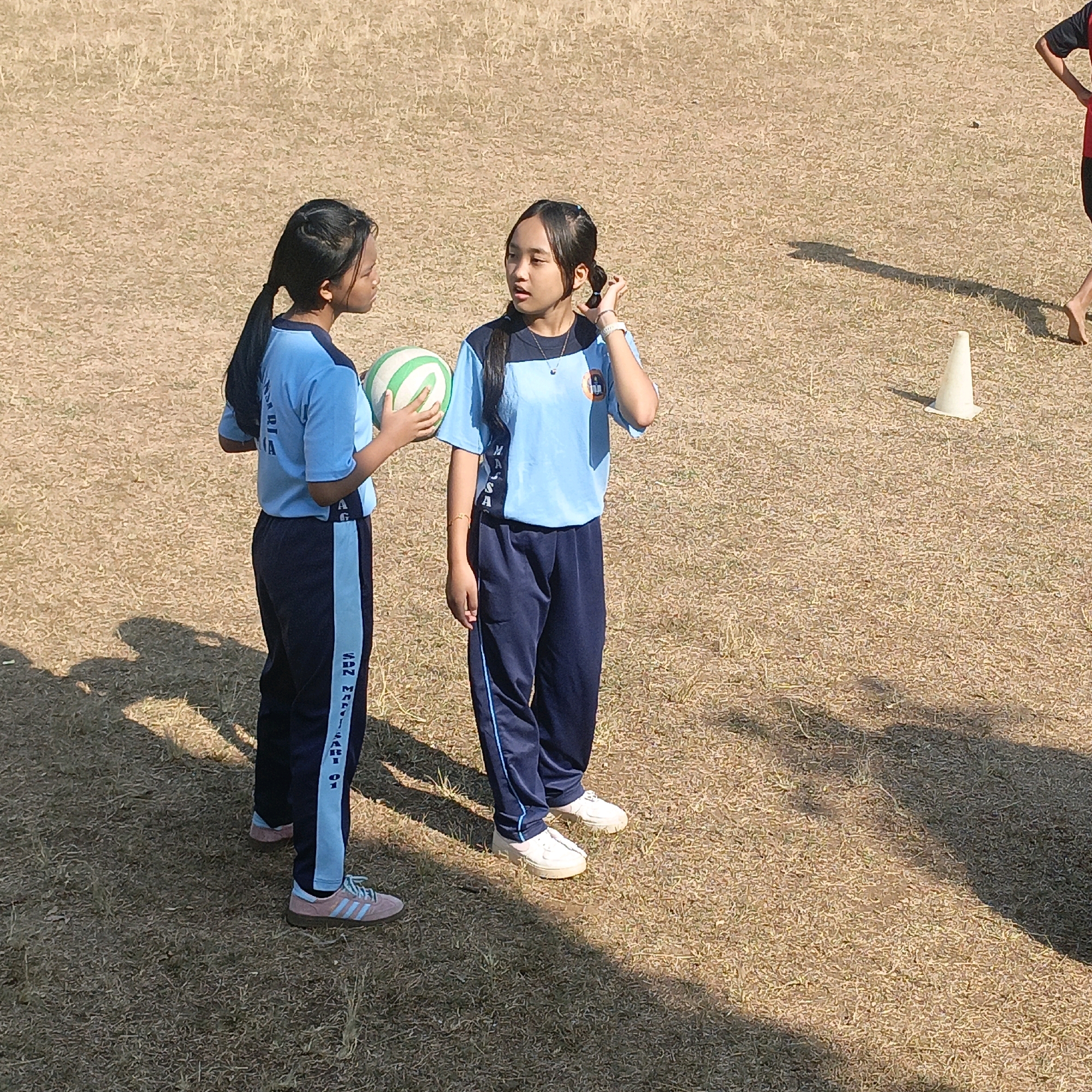 Two young girls in athletic uniforms stand on a dry, grassy field. The girl on the left wears a light blue shirt with dark blue stripes down the sides of her pants and holds a green and white striped ball. The girl on the right wears a similar light blue shirt with a logo on the left chest and dark blue pants. Both girls have dark hair styled in ponytails. They appear to be in conversation. In the background, a white traffic cone is visible, and a shadow of a person is cast on the ground. The scene is likely an outdoor sports field, possibly part of a school or athletic complex. The overall environment suggests a warm, dry climate. The text visible on the pants of the girl on the left reads "SUN MARI -SARI 01". This suggests a possible location related to a school or institution in Salatiga, Indonesia, as "Sari" can be a common element in Indonesian school names, and "01" might refer to a specific branch or year.