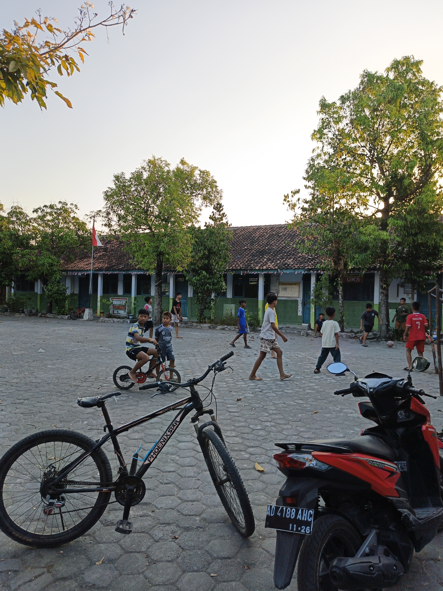 The image depicts a schoolyard in Trucuk, Indonesia, during late afternoon. A group of boys are engaged in various activities. In the foreground, a black bicycle with "PHOENIXSTAR" branding is parked. To the right, a red scooter with license plate "AD 4188 AHC 11.26" is also parked. Several children are playing with a soccer ball in the middle ground, while two younger boys are riding small bicycles. In the background, a building with a red tiled roof, typical of Indonesian architecture, serves as the school. A red and white Indonesian flag is visible flying from a flagpole near the building. The ground is paved with interlocking grey bricks. The scene is bathed in the warm glow of the setting sun, casting long shadows and highlighting the green foliage of the trees scattered throughout the yard.