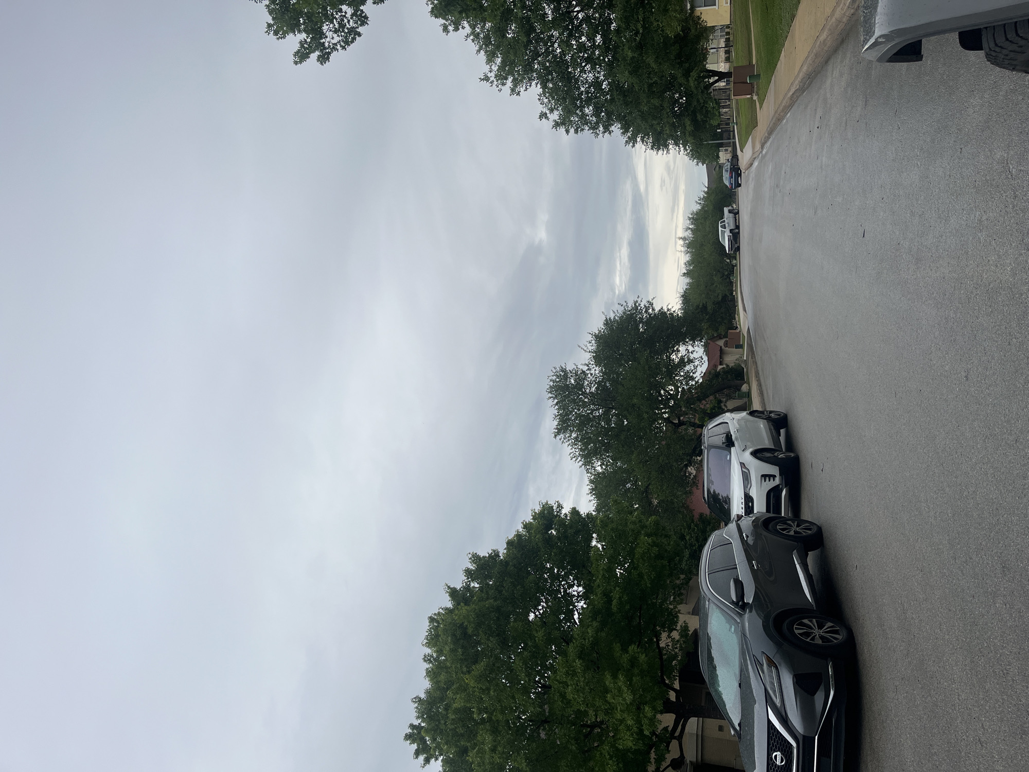 The image depicts a residential street in San Antonio, United States of America. Two cars are parked along the right side of the street, a gray Nissan Murano and a white Land Rover Range Rover. Other vehicles are visible further down the street. The sky is overcast. The road appears wet. Trees line both sides of the street. Houses are visible behind the trees.