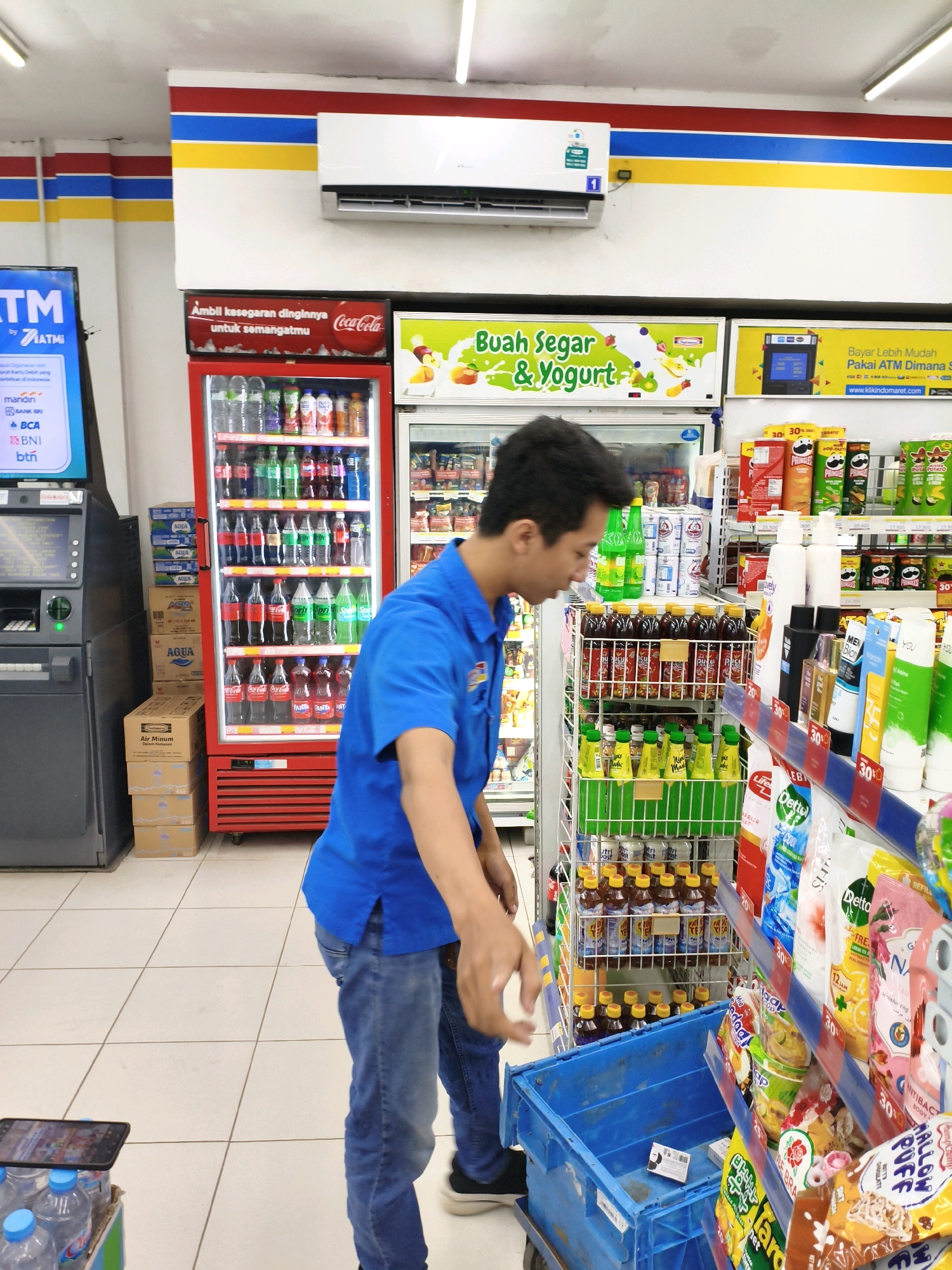 In the city of Balesono, Indonesia, a male convenience store employee wearing a blue polo shirt and blue jeans is stocking shelves. He is standing in an aisle filled with merchandise, facing right, and appears to be reaching for a product on a shelf. To his left, a red cooler stocked with beverages is visible, and to his right, shelves display various packaged goods, including chips, drinks, and cleaning supplies. A blue plastic crate is positioned in front of him, possibly for organizing inventory. An ATM machine is present on the far left side of the image. The store appears to be well-lit with fluorescent lighting, and the walls are decorated with colorful stripes.