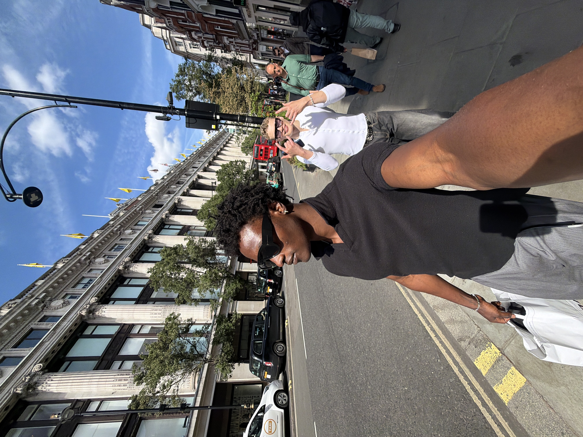 The image depicts a person taking a selfie on a street in London, United Kingdom. The person is wearing sunglasses and holding a shopping bag. Another person is making a hand gesture towards the camera. A red double-decker bus is visible in the background, along with other pedestrians. There are taxis parked on the side of the street, adjacent to a large building with ornate architectural details. The sky is blue with scattered clouds. Street lights and traffic signals are also present.