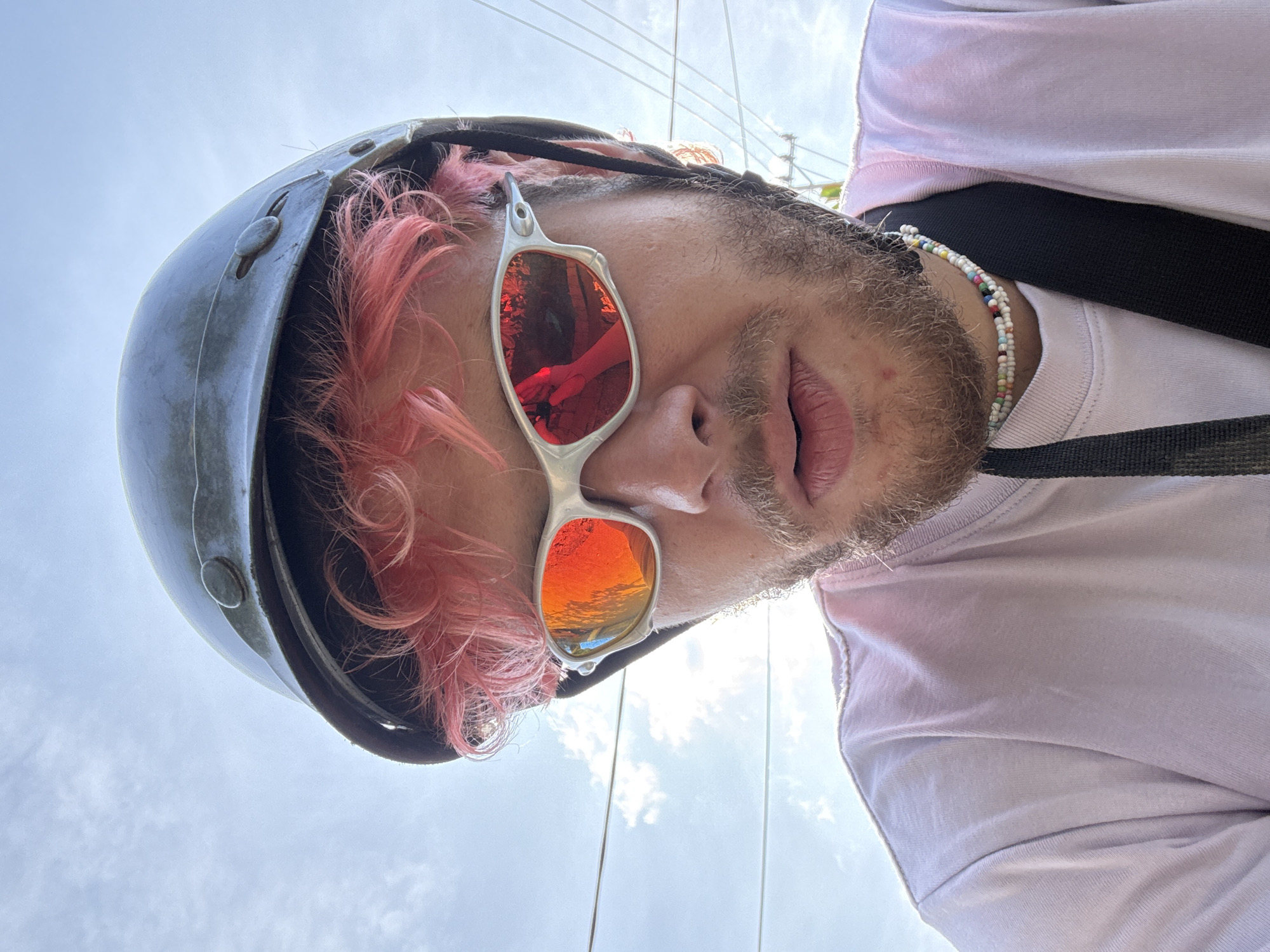 A low-angle, point-of-view shot depicts a person wearing a helmet and sunglasses. The helmet is dark gray, and the hair protruding from underneath the helmet is dyed pink. The sunglasses have silver frames and red lenses, reflecting trees and a hand holding a camera. The person has a beard and is wearing a light pink T-shirt with a black strap across the chest. A beaded necklace is visible around the neck. Above the person are power lines and a cloudy sky. Without further information, it's impossible to confirm the location to be Đà Nẵng, Vietnam.
