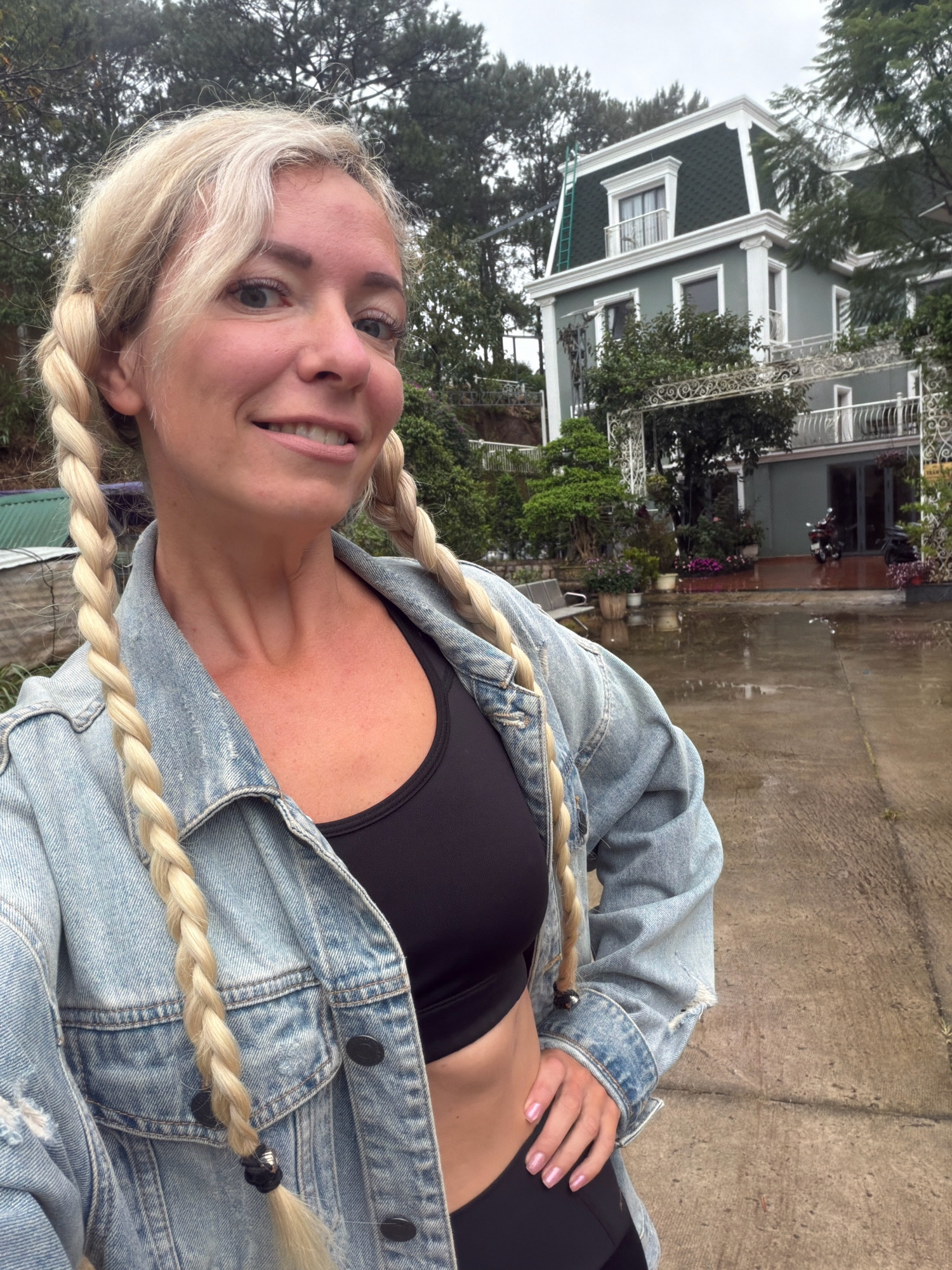 A woman with blonde hair styled in two braids stands in front of a two-story house. The house is painted a light grey and has a dark green mansard roof. The woman is wearing a denim jacket over a black sports bra and black bottoms. She is smiling and has her left hand on her hip. The ground in front of the house is wet and reflects the surroundings, indicating it has recently rained. There are potted plants and trees around the house, and a motorcycle is parked near the entrance. The scene appears to be located in a residential area, possibly a guesthouse or a private home, in Xuân Trường, Đà Lạt, Vietnam. The overall atmosphere is casual and relaxed.
