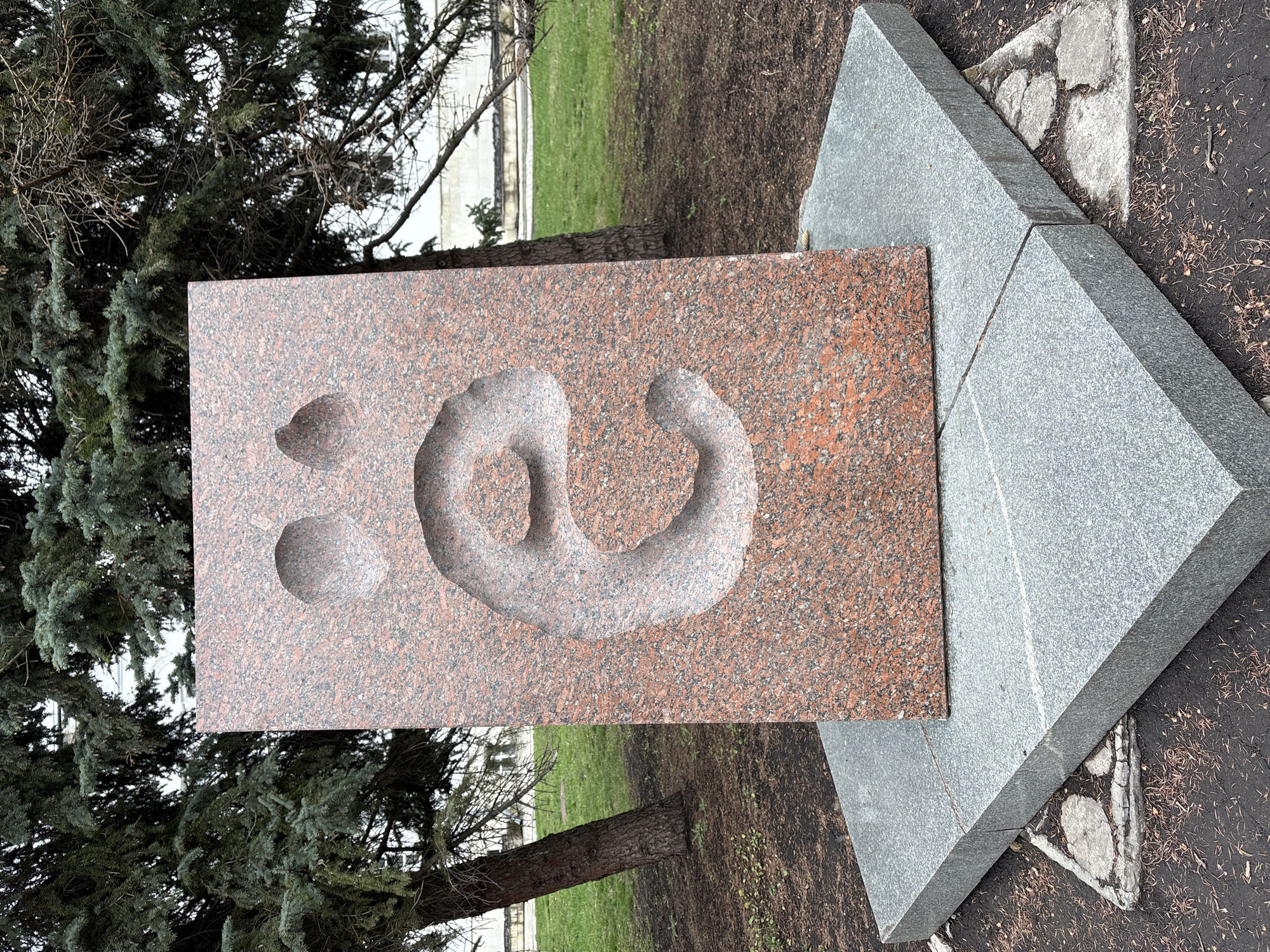 The image shows a red granite sculpture set against a backdrop of a tree and a grassy area. The sculpture features a relief carving of what appears to be a human hand forming a "shisha" gesture, with two additional circular depressions. The granite slab is mounted on a gray, pyramidal base. The surrounding environment suggests an outdoor setting, possibly a park or public space. Based on available context, this image most likely depicts the "Monument to Ulyanovsk Drivers," a public artwork located in Ulyanovsk, Russia.