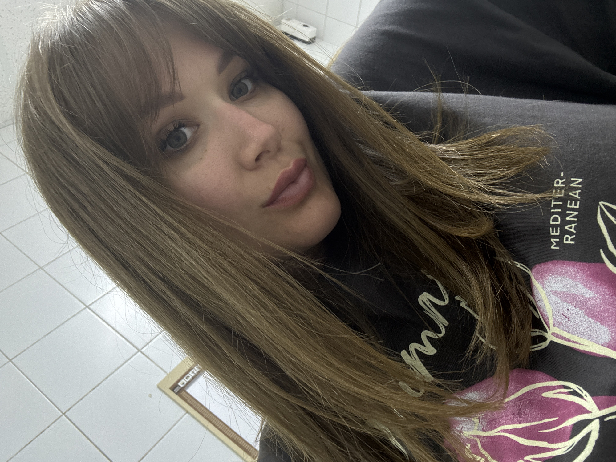 A woman with light brown hair and bangs is posing for a selfie. She is wearing a black shirt with floral designs and text that includes "MEDITERRANEAN". The background consists of white tiles on the wall and floor. The location in the city of Steblitskii, Russia is not discernible from this image.