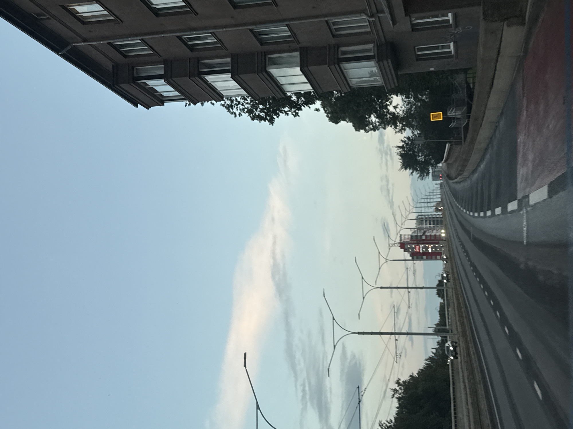 The image shows a view from a vehicle on a multi-lane road in Tallinn, Estonia. The road curves to the right, with a dark, multi-story apartment building on the right side of the frame. The building features a bay window structure on its upper floors. On the left side of the road, there is a median with trees and several streetlights with distinctive curved arms. Further down the road, there are more streetlights and some buildings in the distance, one of which is red and appears to be a modern structure with lit windows. The sky is light blue with some wispy clouds, suggesting it is either early morning or late evening. There are a few cars visible on the road, with their headlights on. A yellow traffic sign with a pedestrian icon is visible near the building on the right.
