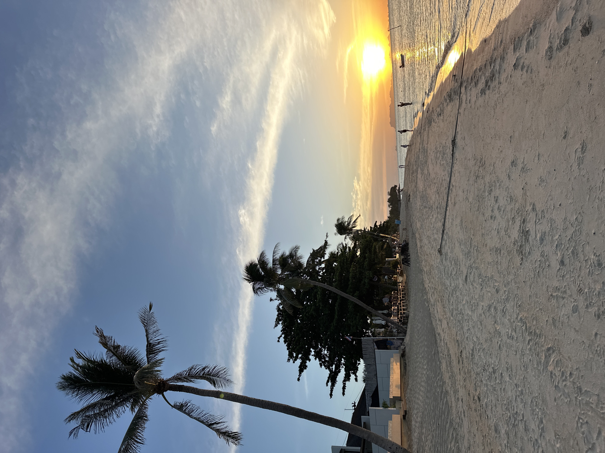 A wide-angle, eye-level shot captures a serene sunset scene on a beach in the Ko Samui District of Thailand. The sun, a bright, glowing orb, is setting on the horizon, casting a warm, golden light across the sky and the calm ocean. Wispy clouds streak across the blue sky, tinged with orange and yellow from the sunset.
On the beach, the sand is a light beige color, marked with the imprints of footprints and subtle ripples from the receding tide. To the left, a tall, slender palm tree with fronds reaching upwards dominates the foreground. Behind it, a row of buildings with white walls and visible awnings are situated. Further down the beach, a line of darker green trees, including more palm trees, creates a lush backdrop. Outdoor seating areas with tables and chairs are visible under some of the trees, suggesting a restaurant or bar.
In the middle ground, several people are scattered along the shoreline, some standing in the shallow water, others walking. Their silhouettes are outlined against the bright sunset. A small boat is anchored in the water near the horizon. The overall atmosphere is peaceful and tranquil, indicative of a beautiful tropical evening.