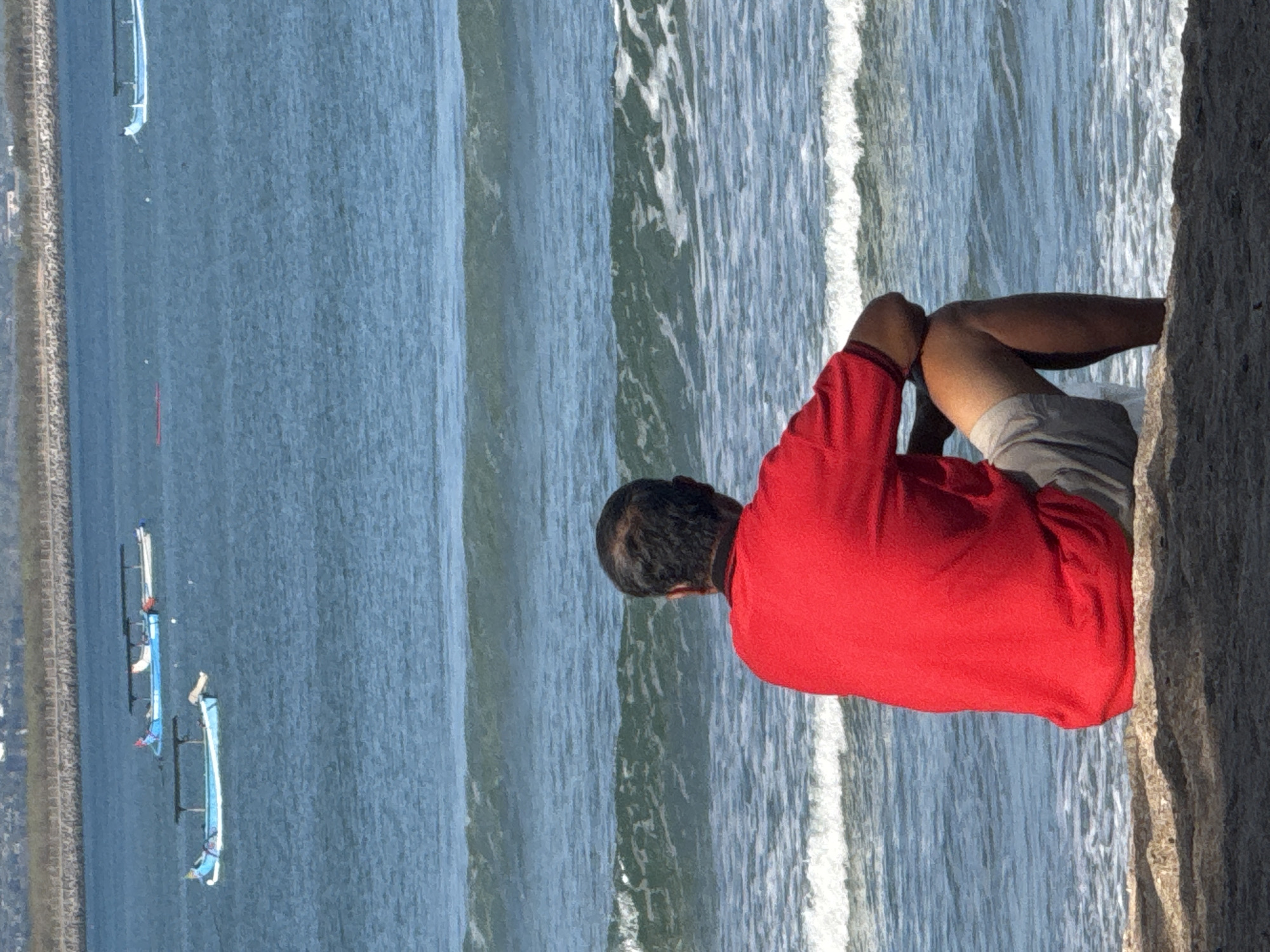A man wearing a red shirt and gray shorts sits on a sandy shore, facing the ocean. The man is positioned on the left side of the frame, with his back to the viewer, his arms resting on his knees. The ocean stretches out before him, with small waves breaking on the shore. In the distance, several traditional Indonesian outrigger boats, known as jukungs, are visible on the water. The background shows a coastline with some vegetation and structures, indicative of a developed area. This scene is typical of the Kuta beach area in Bali, Indonesia, known for its popular surf breaks and vibrant beachfront activity.