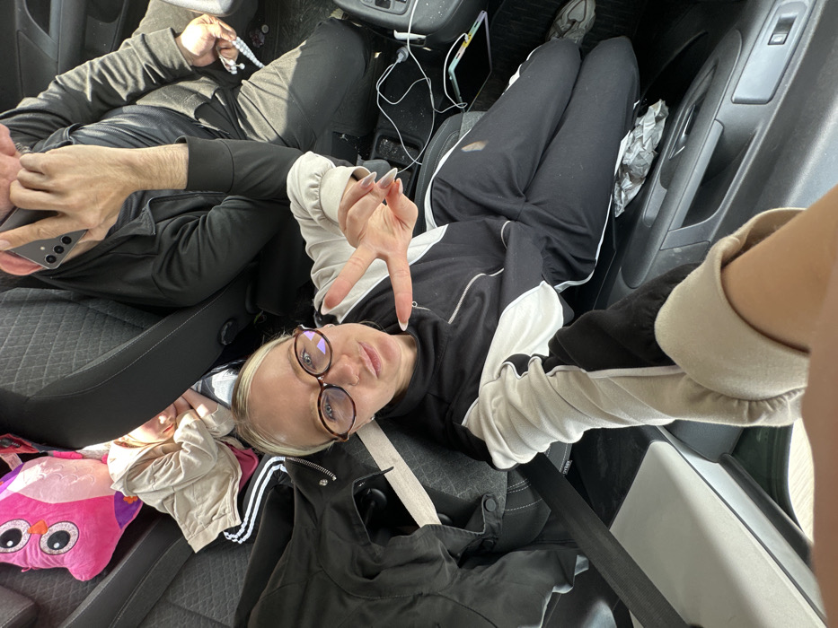 Inside a vehicle in Torbalı, Turkey, three individuals are present. In the foreground, a blonde-haired female, wearing glasses and a black and white tracksuit, looks directly at the camera, forming a peace sign with her left hand. Her right arm and lower body are partially visible. To her right, a male figure in the front seat is on a phone call, holding a light-colored smartphone to his right ear, while his left hand holds a white string of prayer beads. To the female's left, a child wearing a light-colored jacket is partially visible, appearing to rest against a dark seat, with a pink owl-shaped plush toy situated behind them. A black jacket is draped over the seat next to the female. The vehicle's interior features dark fabric seats, visible seatbelts, and a center console with cables, a dark smartphone, and a crumpled wrapper. The floor of the vehicle contains some visible debris.