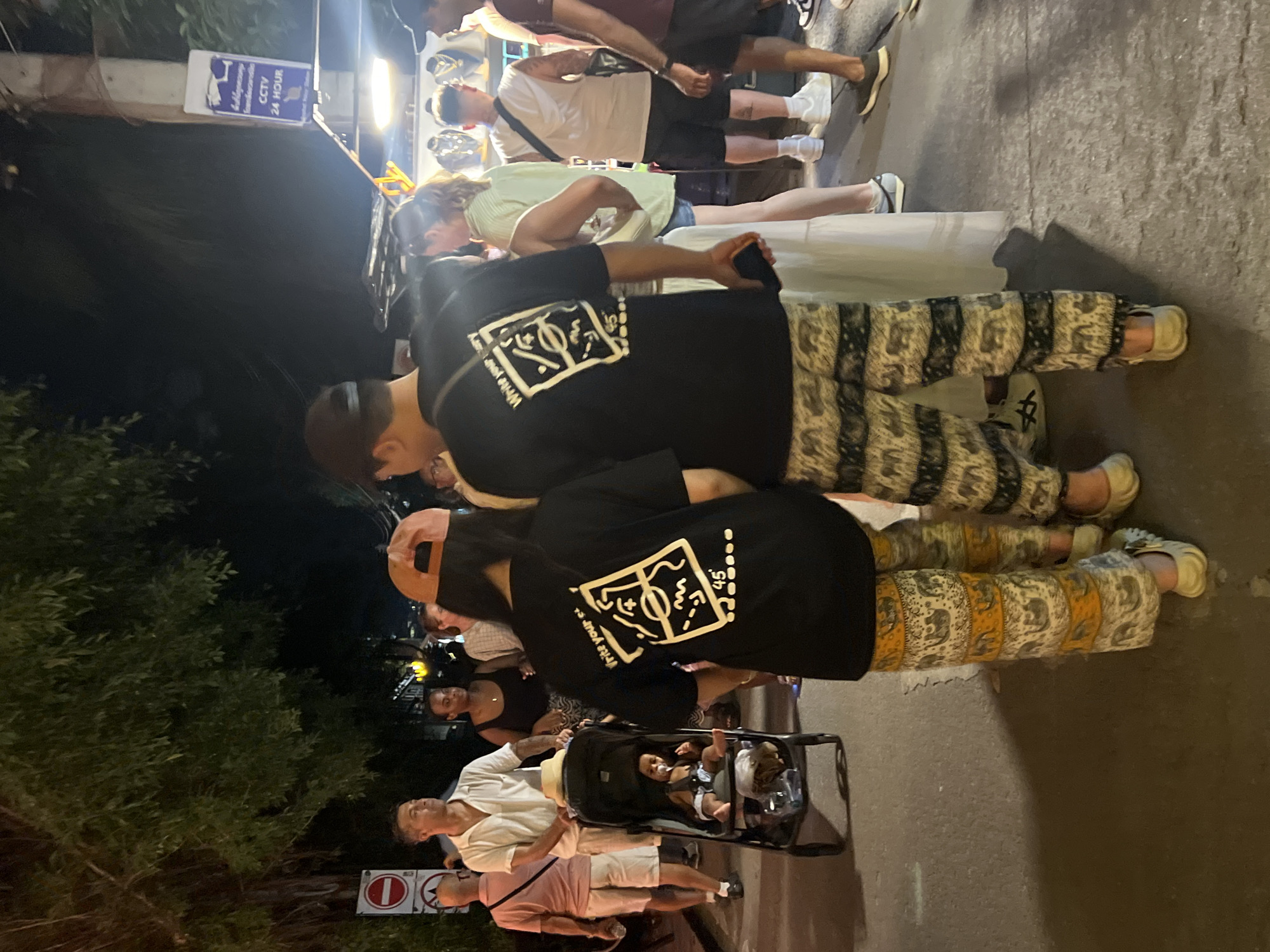 A nighttime shot shows a crowded street scene. People are walking, standing in groups, and pushing strollers. The lighting is a mix of ambient streetlights and brighter illumination from what appears to be street vendors. One person is wearing a black shirt with a design on the back, and they are next to someone wearing a baseball cap. There are shops and signs visible in the background, including one that reads "CCTV 24 Hour". The scene seems to be in a busy urban area.