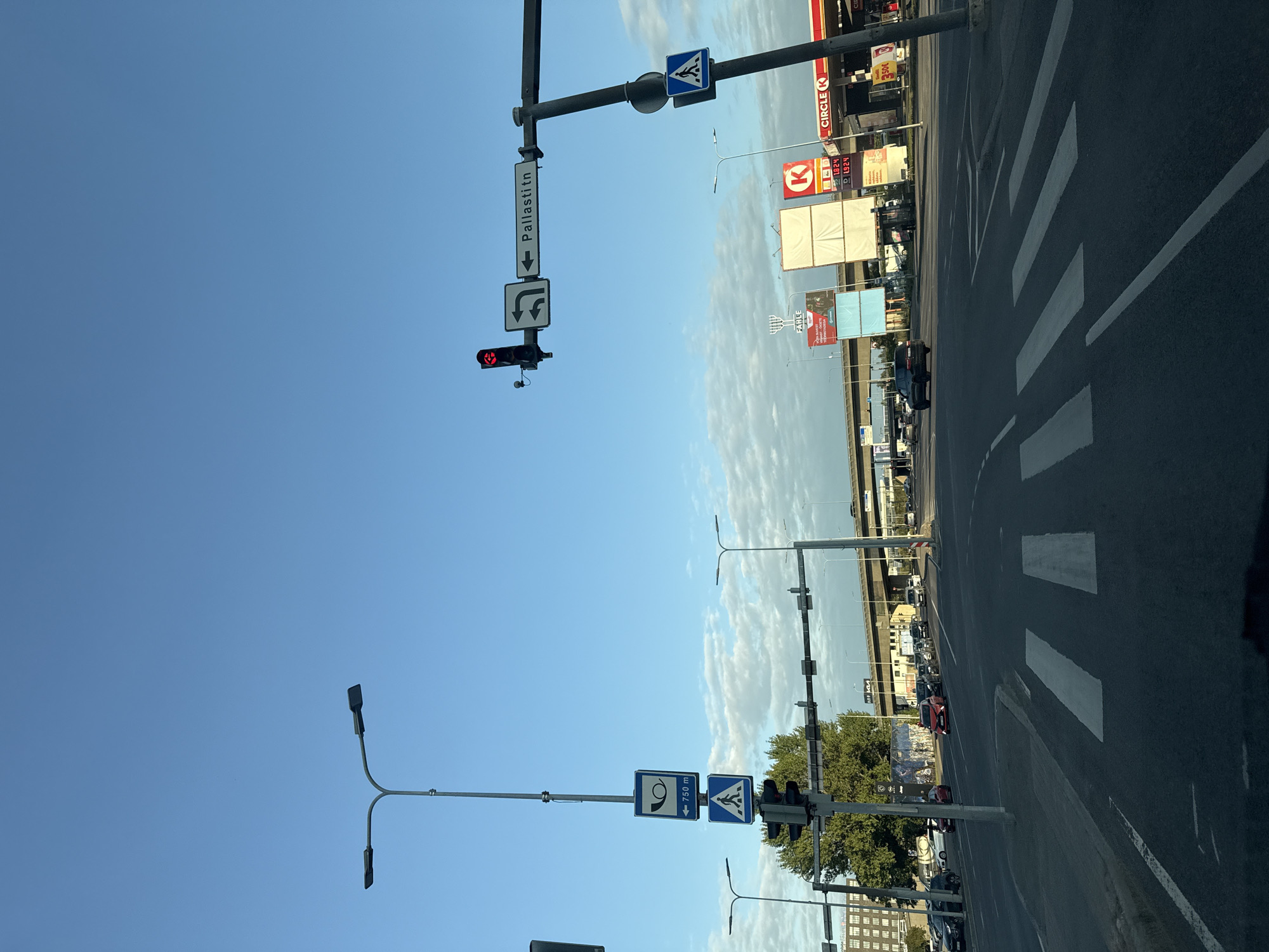 The image shows a street scene in Tallinn, Estonia, at an intersection. The sky is clear and blue with some scattered clouds. In the foreground, there is a road with a crosswalk. To the left, a tall streetlamp with two lights stands beside a traffic light pole with a pedestrian crossing sign. Further down the street, trees and buildings are visible. To the right, a traffic light with a red signal hangs above a street sign indicating "Pallasti tn" with arrows for turning left and going straight. There's also a pedestrian crossing sign on this side. In the background, an elevated highway and a Circle K gas station with advertising billboards can be seen. Several cars are on the road, some moving and some stopped. The overall impression is that of a typical urban intersection.