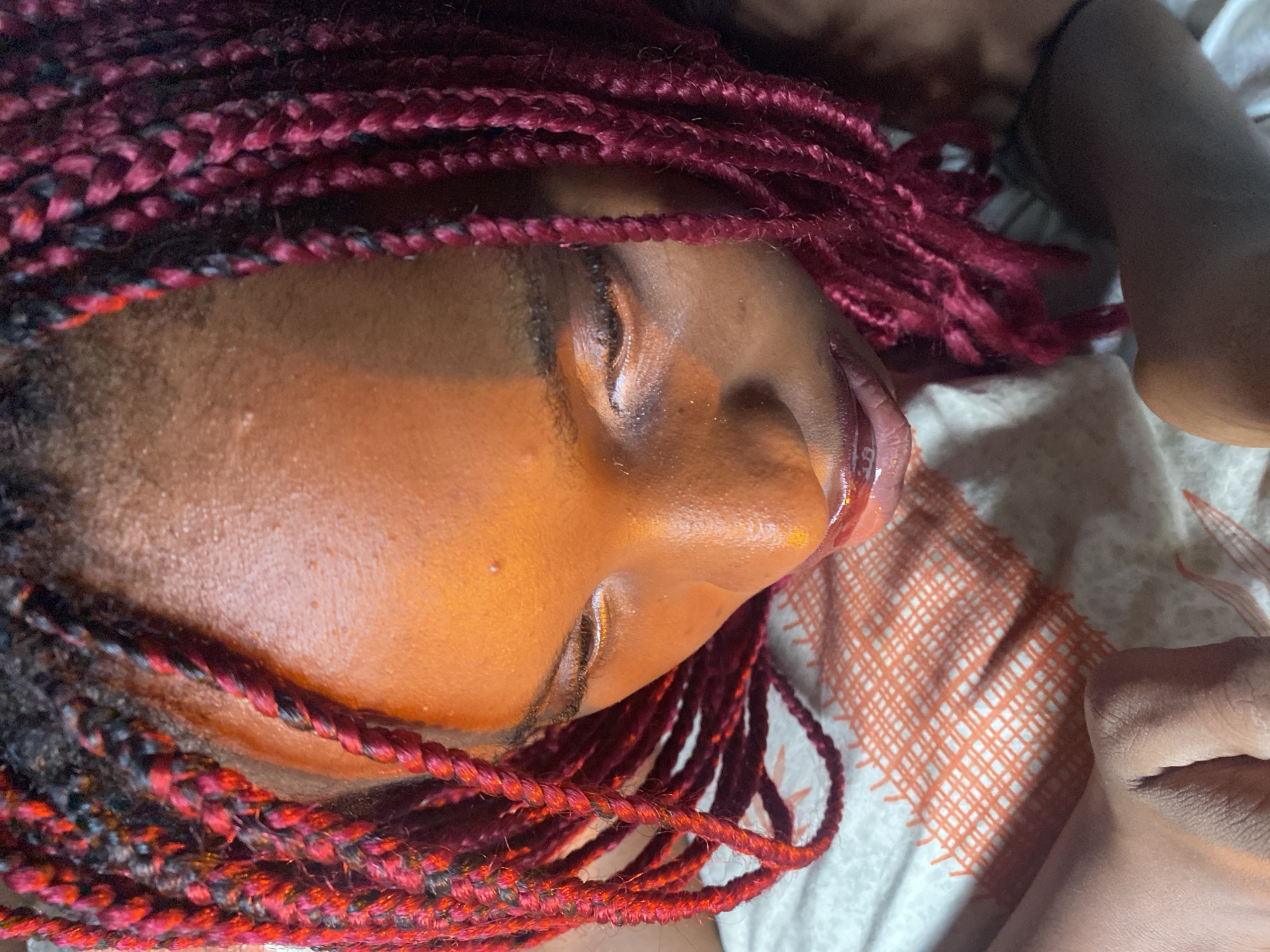 The image is an extreme close-up, overhead shot of a young Black person with braided hair that is a deep red color. The person's face is illuminated with an orange light, casting shadows and highlighting the skin's texture. Their eyes are closed, and their head is tilted downwards. The person is lying on a patterned surface, possibly a bed or a blanket, with a repeating orange grid design. A hand is visible in the lower left of the frame, resting on the patterned surface. The overall impression is an intimate and artistic portrait, with a focus on light and texture. The scene is not identifiable as being in Iranla, Nigeria, or any other specific location.