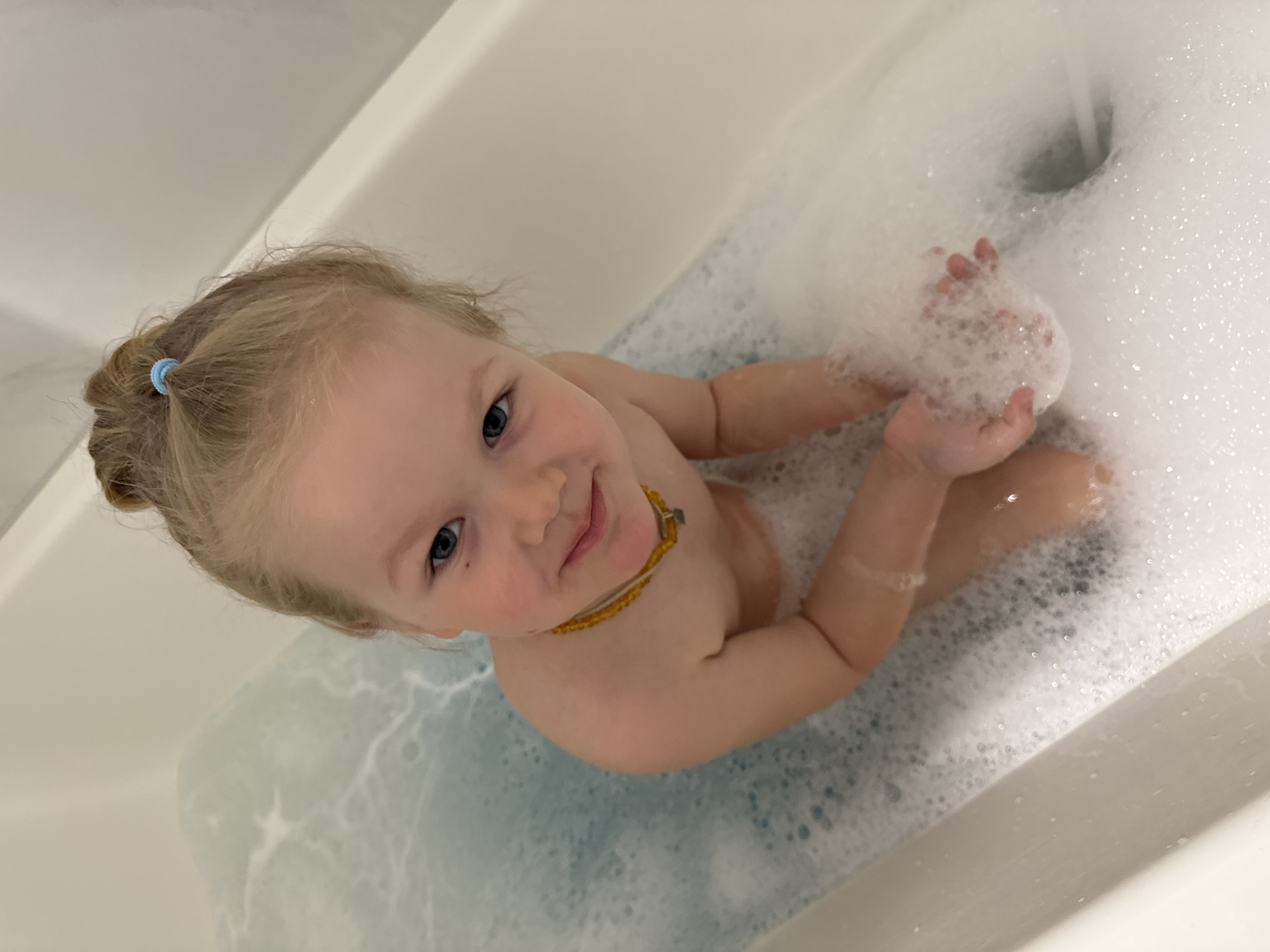 A young, light-skinned child with blonde hair pulled back with a blue hair tie is sitting in a white bathtub filled with blue-tinted water and copious white bubbles. The child is holding a handful of bubbles, looking up and smiling slightly at the camera. The setting appears to be a domestic bathroom. The city is Ферма, Russia.