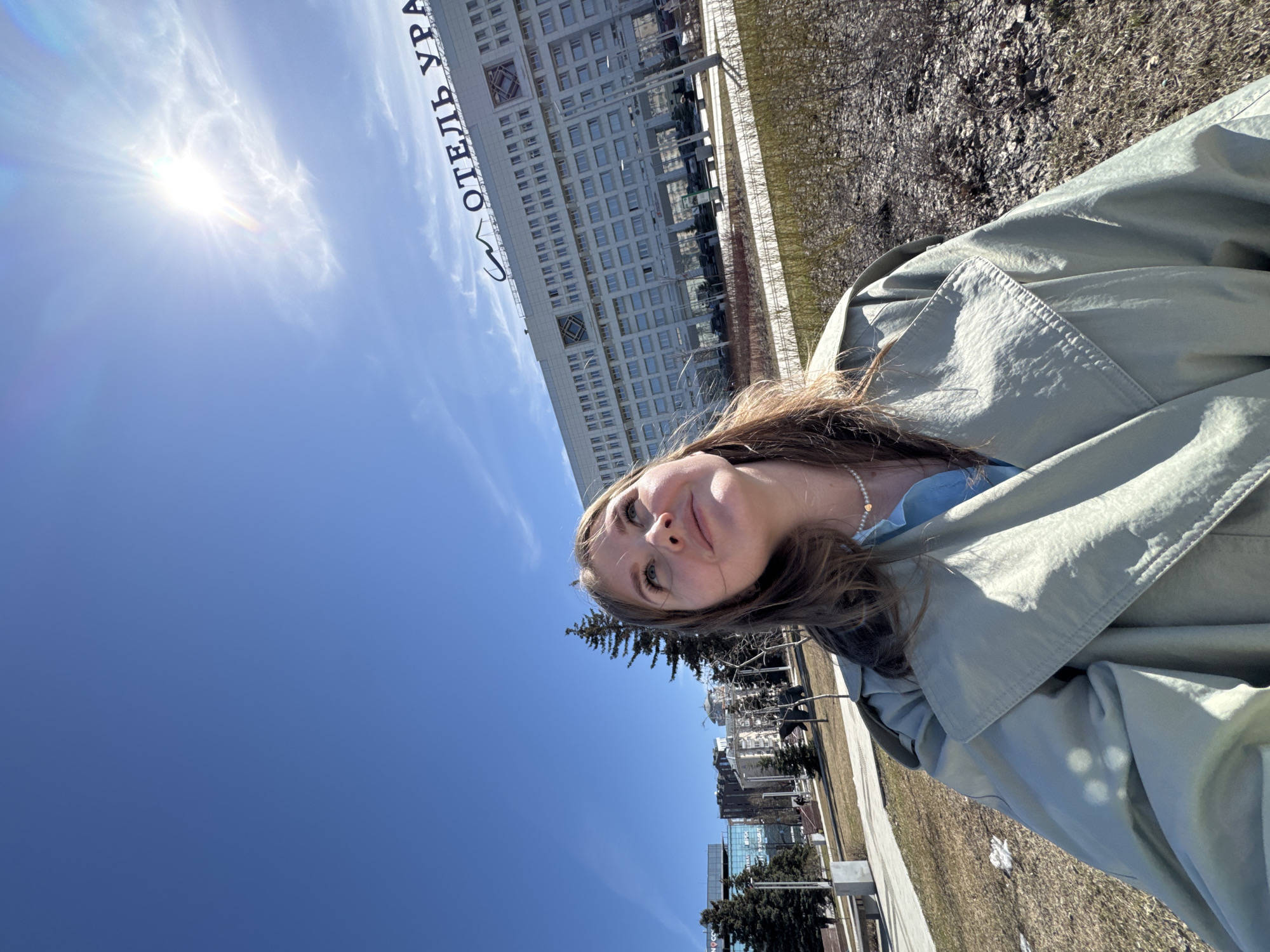 A young woman poses for a selfie outside the Hotel Ural in Perm, Russia. The Hotel Ural's sign is visible in the background. The sky is clear blue with a bright sun. The ground is covered with dry grass and fallen leaves. Other buildings and trees are also in the background, lining a pedestrian walkway with benches and light poles. She is wearing a light-colored trench coat and a delicate pearl necklace.
