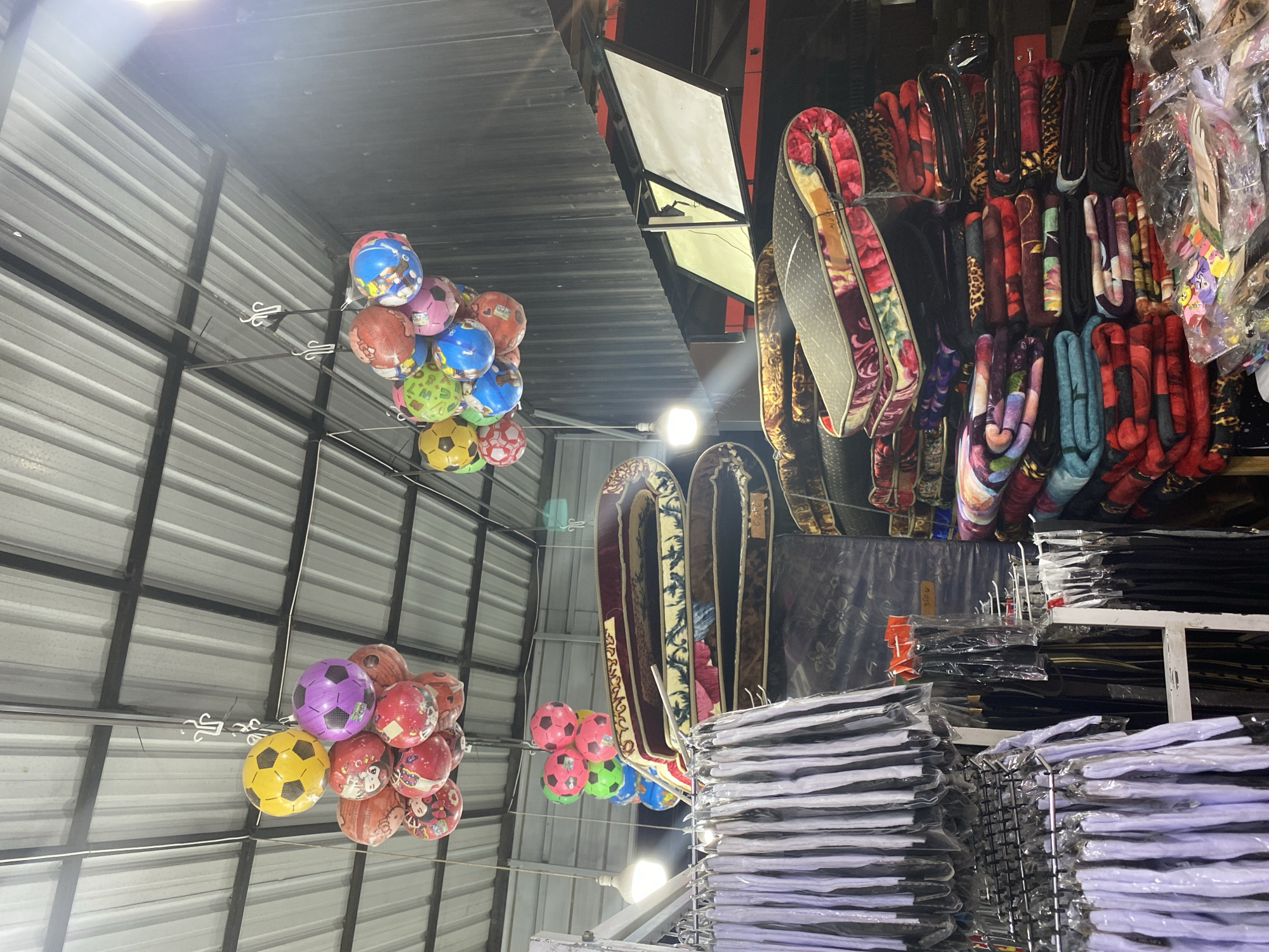 In Lamteumen Barat, Indonesia, a market stall displays a variety of goods suspended from the corrugated metal ceiling. Clusters of colorful inflatable balls, some designed to resemble soccer balls, hang overhead, adding a playful element to the scene. Below, neatly stacked mattresses and blankets, adorned with intricate floral and animal prints, are arranged for sale. To the left, rows of apparel, possibly uniforms or clothing items, are hung on racks, encased in clear plastic packaging. The stall appears to be well-stocked, with a wide selection of textiles and decorative items. The lighting is bright, suggesting an indoor setting, possibly a covered market or a shop.