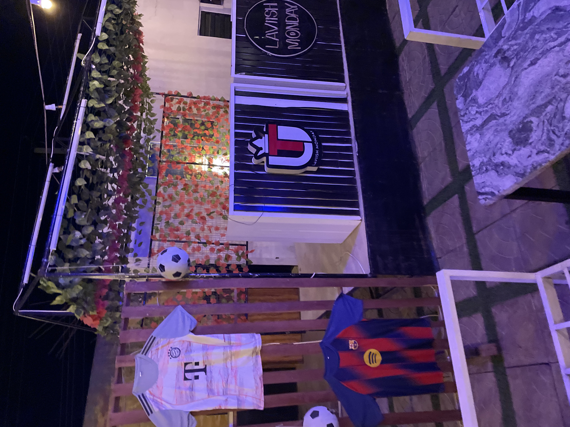 This image depicts an outdoor or semi-outdoor establishment, likely a cafe, bar, or lounge, in Jos, Nigeria, at night. The scene is illuminated by artificial lights, casting warm yellow and cool purplish hues.
Dominating the left side is a dense arrangement of green faux ivy and red/orange foliage, hanging from an unseen support structure. Below this, a wooden slatted fence or railing is used to display sports memorabilia. Two soccer (football) jerseys are prominently featured: one on the left, predominantly white with yellow and grey patterns, bearing a large black "T" logo (associated with Bayern Munich and T-Mobile), and another on the right, a red and blue striped jersey with a yellow circular logo (associated with FC Barcelona and Spotify). A classic black and white soccer ball rests on the wooden structure near the white jersey, with another partially visible further right.
Above the foliage, two distinct signs for "LAVISH MONDAY" are mounted on a dark, horizontally slatted facade. The upper sign is a circular black emblem with "LAVISH MONDAY" inscribed in white. Below it, a stylized white "L" with a prominent red "T" forms a logo, also bearing the text "LAVISH MONDAY" in smaller font.
To the far right, a white table with a grey, marble-patterned top is visible, resting on what appears to be a tiled or paved ground. The overall setting suggests a casual and inviting atmosphere, potentially a popular spot for football enthusiasts.
