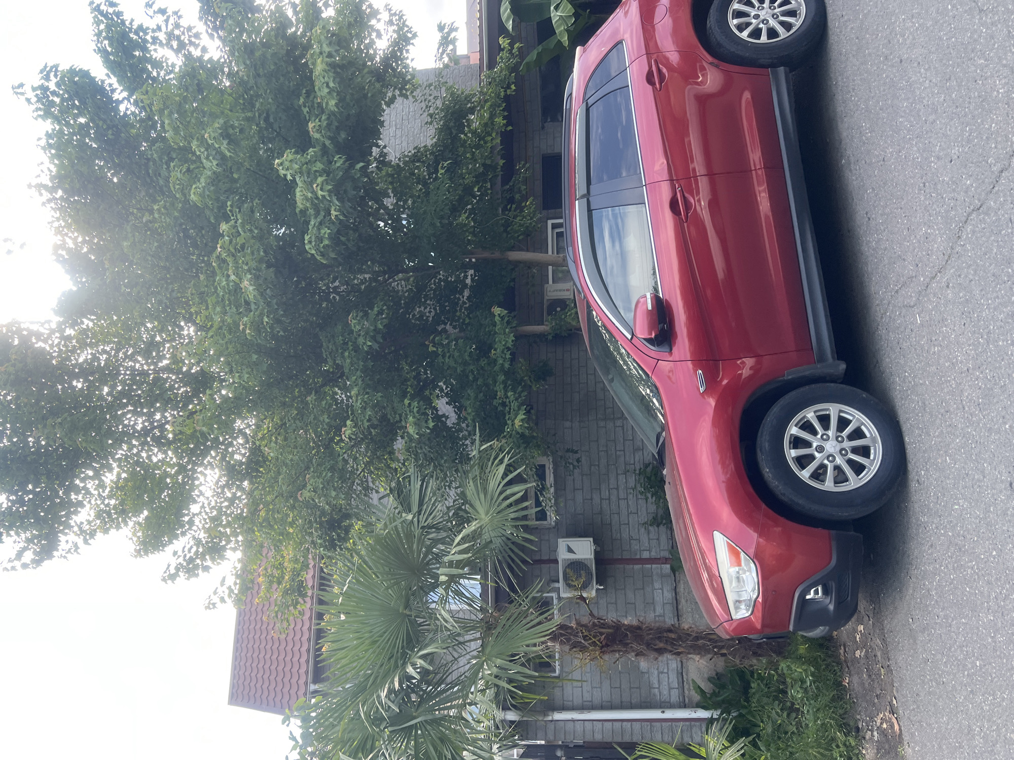 A red Mitsubishi SUV is parked on a street in front of a building. The building appears to be a house with a brick exterior and a red tiled roof. There are trees and a palm tree in front of the house. An air conditioning unit is mounted on the exterior wall of the building. The sky is overcast with some sunlight filtering through the trees. The image is taken from a slightly low angle, looking up at the house and trees.