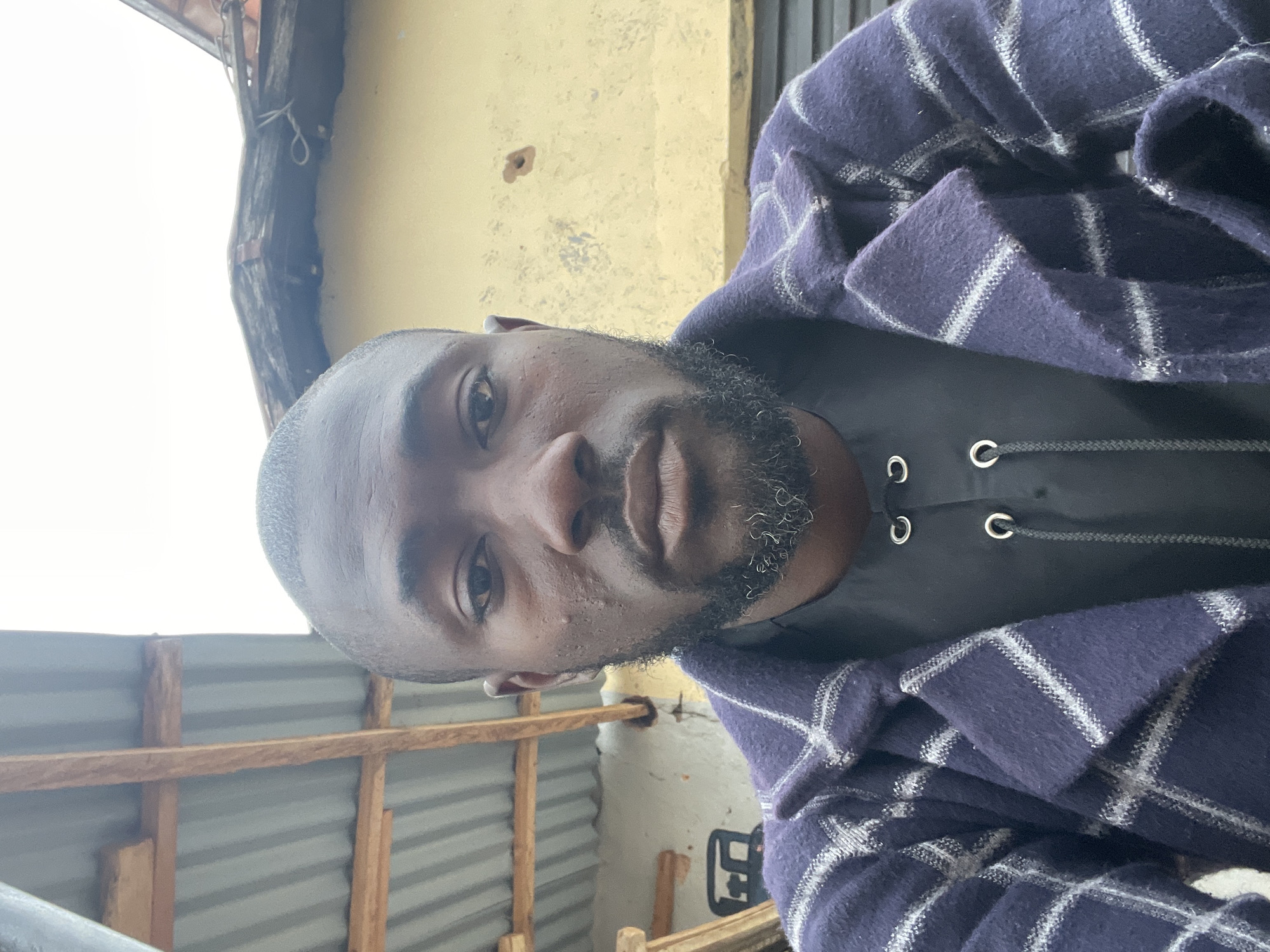 The image is a selfie taken by a man in his late 20s or early 30s, with dark skin, a short-cropped haircut, and a beard. He is wearing a dark, possibly black, shirt with decorative eyelets and lacing, and a navy blue and white plaid jacket. The background appears to be outdoors, with a corrugated metal roof structure visible above and to the left. To the right, a pale yellow wall with some texture and a small dark mark is visible. The overall setting suggests an urban environment in Jos, Nigeria, likely in a residential or informal commercial area, perhaps during daylight hours given the lighting.