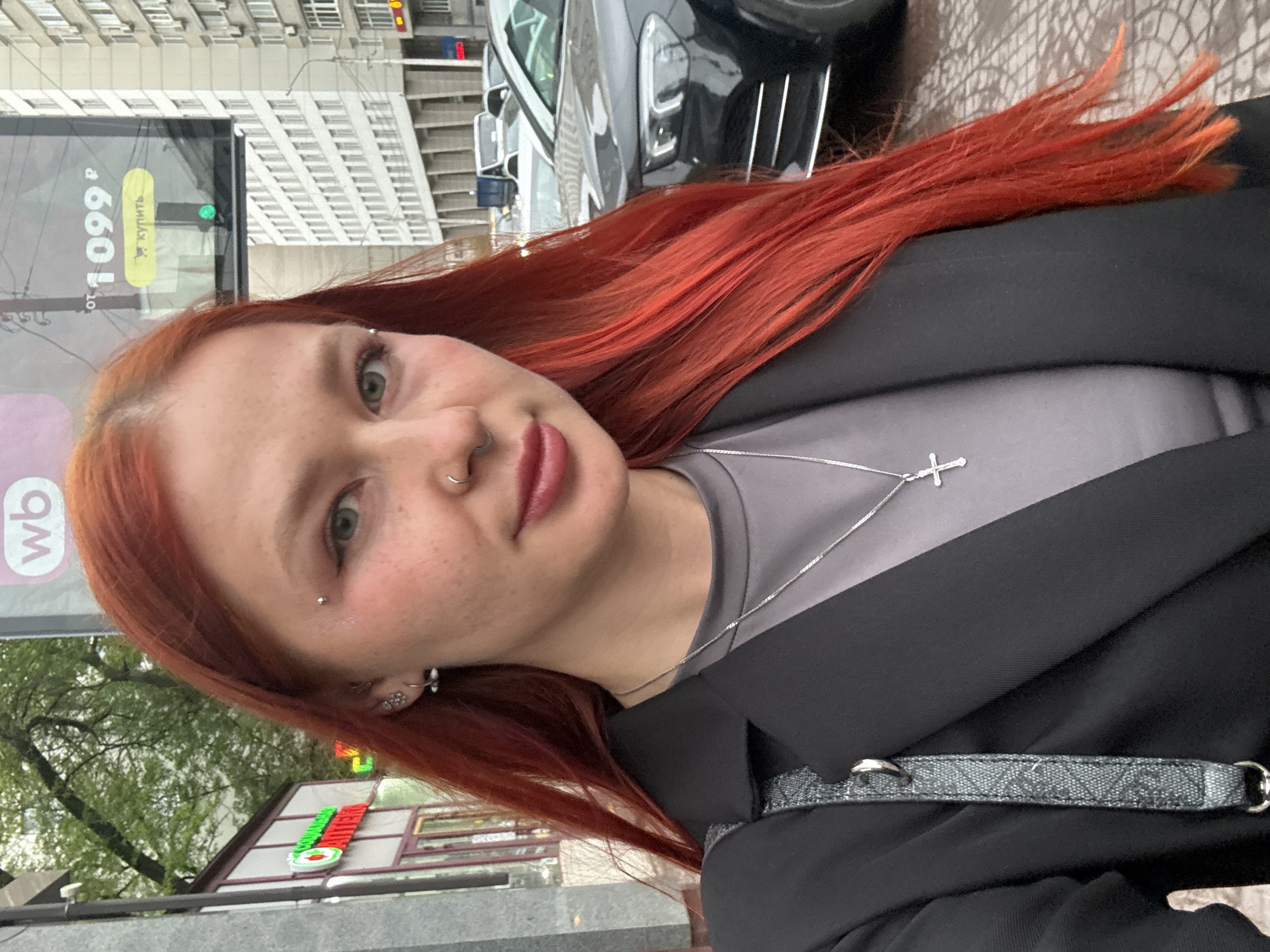 A selfie captured on a city street in Rostov-on-Don, Russia. The subject is a fair-skinned person with freckles and vibrant red hair. They have multiple piercings, including a nose ring, eyebrow piercing, and ear piercings. The person is wearing a gray top and a black jacket. A silver necklace with a cross pendant adorns their neck.
In the background, one can see a building and a parked black car. A bus stop sign is visible, displaying the price "1099 p" and a green light. A building with a green and red logo can also be observed. The environment suggests an urban setting.