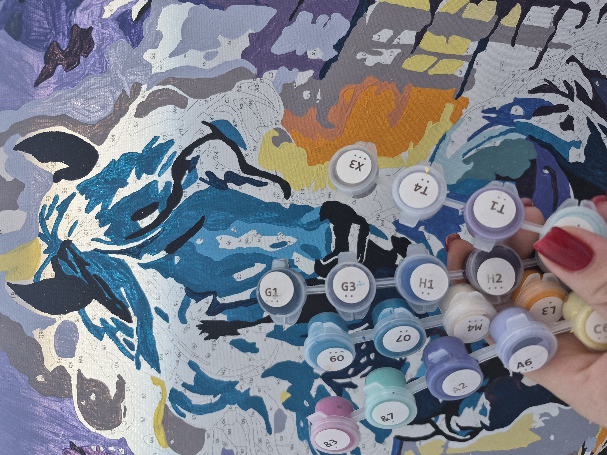 The image shows a partially completed paint-by-numbers canvas and a hand holding small pots of acrylic paint. The canvas depicts an abstract design with various shapes and lines labeled with numbers and letters, indicating the corresponding colors to be applied. The painting is primarily filled with blues, grays, oranges, and yellows. The hand, with a visible red nail polish, holds the paint pots close to the canvas, suggesting an ongoing painting process. The paint pots have white caps with alphanumeric codes matching the canvas labels. The scene is indoors, likely in an artist's studio or hobby space. There is no visible evidence indicating the location is Tobolsk, Russia.