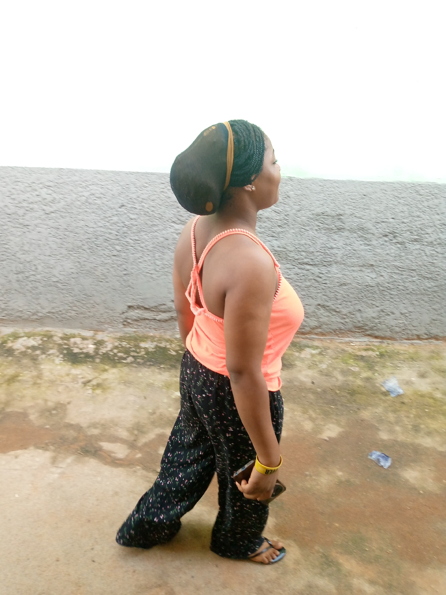 A young Black woman with dark braided hair and a yellow headband is walking outdoors, viewed from the side. She is wearing a coral-colored tank top with beaded straps and black, wide-leg pants with a floral print. She has a yellow bracelet on her left wrist and is holding a dark-colored mobile phone in her right hand. She is wearing blue flip-flops. The woman is walking on a concrete pathway. Behind her is a textured gray wall, and beyond that, a body of water with a rippled surface. The sky above is overcast and white. The ground is a mix of concrete and some patchy green vegetation. Two pieces of clear plastic litter are visible on the ground to the right of the woman. The scene appears to be located in Jauro Yinu, Nigeria, by the water.