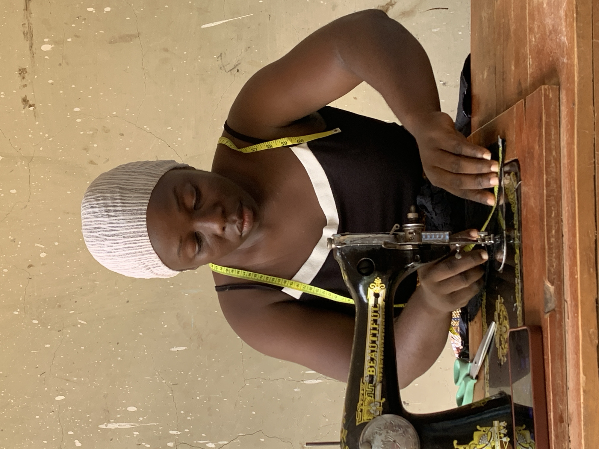 A Black woman, wearing a light pink striped headwrap and a black tank top with white trim, is seated at a wooden table, operating a vintage sewing machine. A yellow measuring tape is draped around her neck. Her hands are engaged with the fabric and the sewing machine. To the left of the sewing machine, a pair of green scissors and a smartphone are visible on the table. The background is a textured, light-colored wall with white splatters. This scene is set in Akwanga, Nigeria.