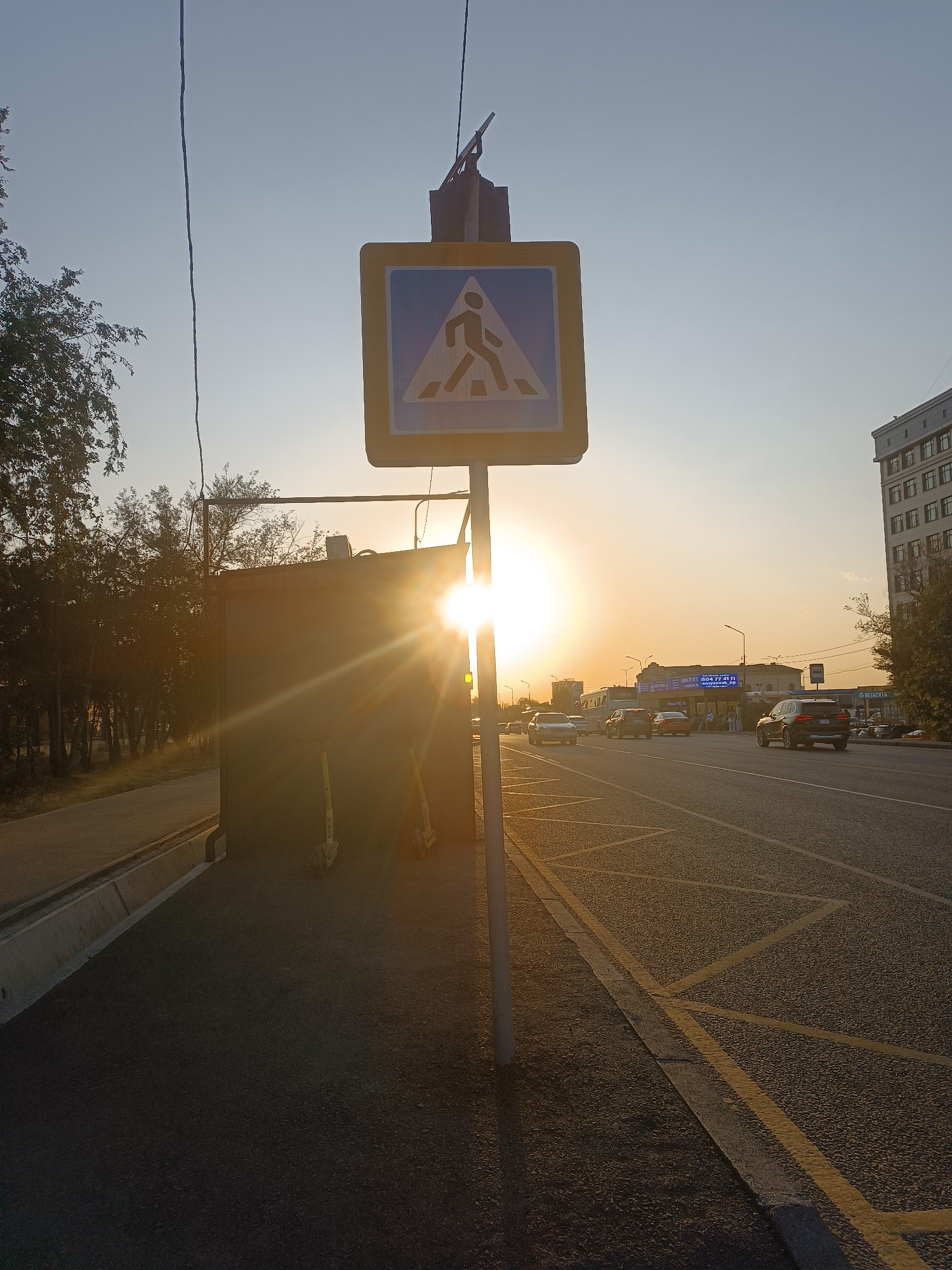 This is a street-level, eye-level, medium shot photograph of a pedestrian crossing sign at sunset in Bishkek, Kyrgyzstan. The sun is low in the sky, casting a warm glow over the scene and creating lens flare. The pedestrian crossing sign is a blue square with a yellow border and a white silhouette of a person walking. A thin, dark electrical wire runs vertically through the upper left portion of the image. To the left of the sign is a dark, rectangular structure, possibly a utility box, with two yellow scooters parked beside it. Beyond the structure, a sidewalk and trees line the street. The road in the foreground is paved, with yellow lane markings visible. Several cars are driving on the road in the middle ground, and a multi-story building is visible on the right side of the frame. The overall impression is a typical urban street scene during the golden hour.