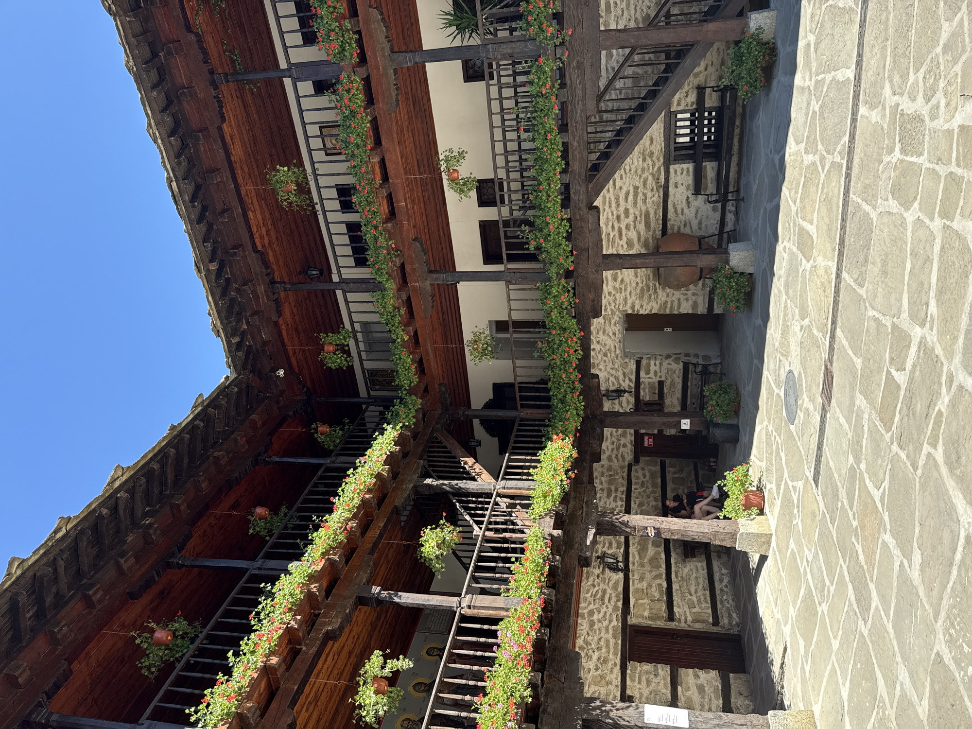 The image shows the interior courtyard of the Troyan Monastery in Oreshak, Bulgaria. The monastery features traditional Bulgarian Revival architecture with two-story wooden galleries surrounding the central courtyard. The galleries are adorned with numerous hanging planters overflowing with red and pink flowers, as well as green foliage. The walls of the lower level are constructed of stone, with wooden doors and windows. A wooden staircase leads up to the second-floor gallery. The courtyard is paved with large, irregular stone slabs. In the courtyard, there are several wooden benches, a large clay amphora, and a few potted plants. Two individuals are seated on a bench on the left side of the courtyard, and another person is partially visible on a bench in the background. The sky above is clear and blue.