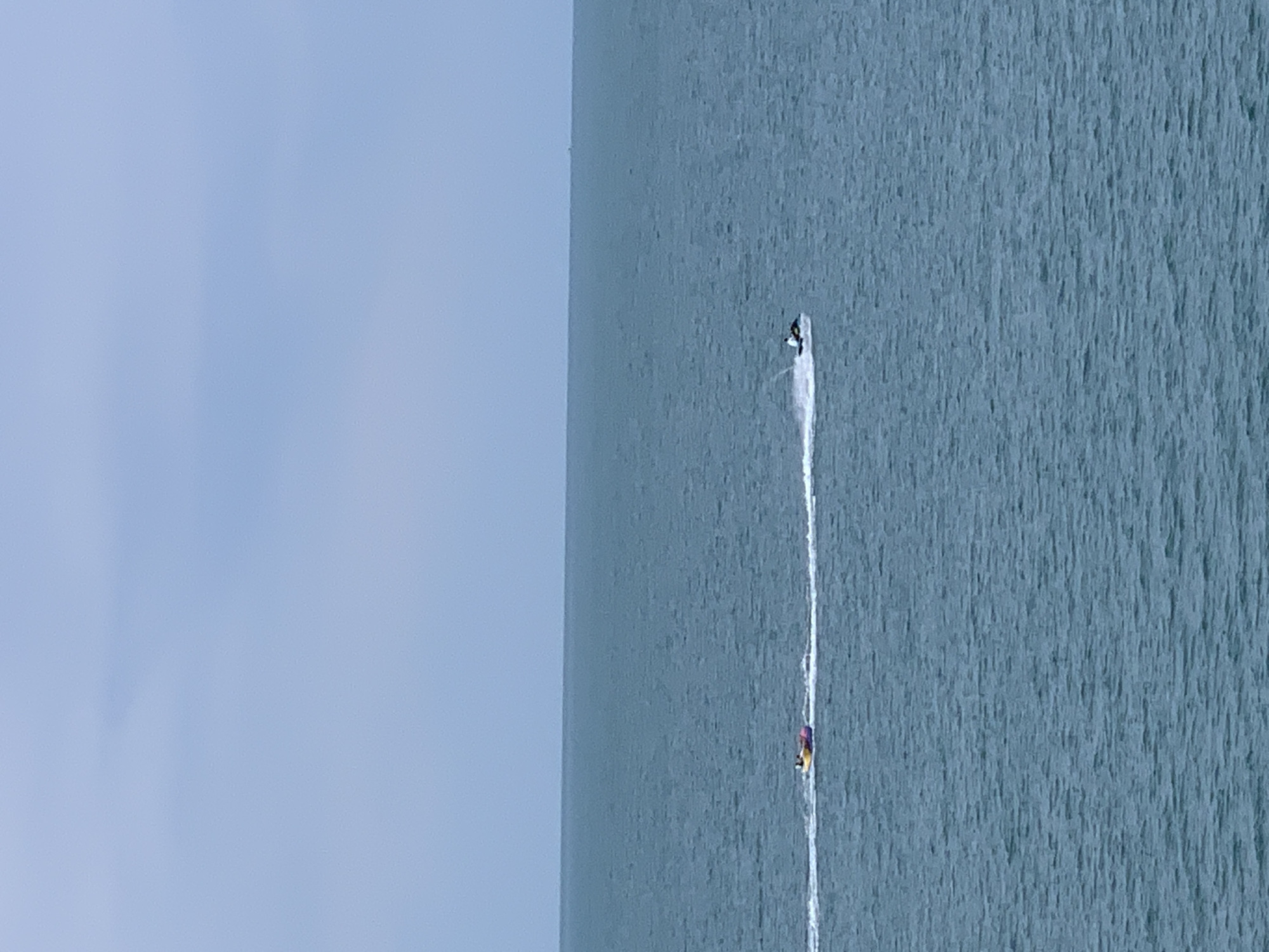 An aerial view shows a vast expanse of turquoise water under a pale blue sky. Two watercraft are visible in the distance. On the left, a yellow and purple inflatable tube is being towed by an unseen boat, leaving a white wake. On the right, a jet ski with a person riding it creates a significant splash. The horizon line is clearly defined, separating the ocean from the sky. The overall scene depicts a recreational activity on a body of water.
