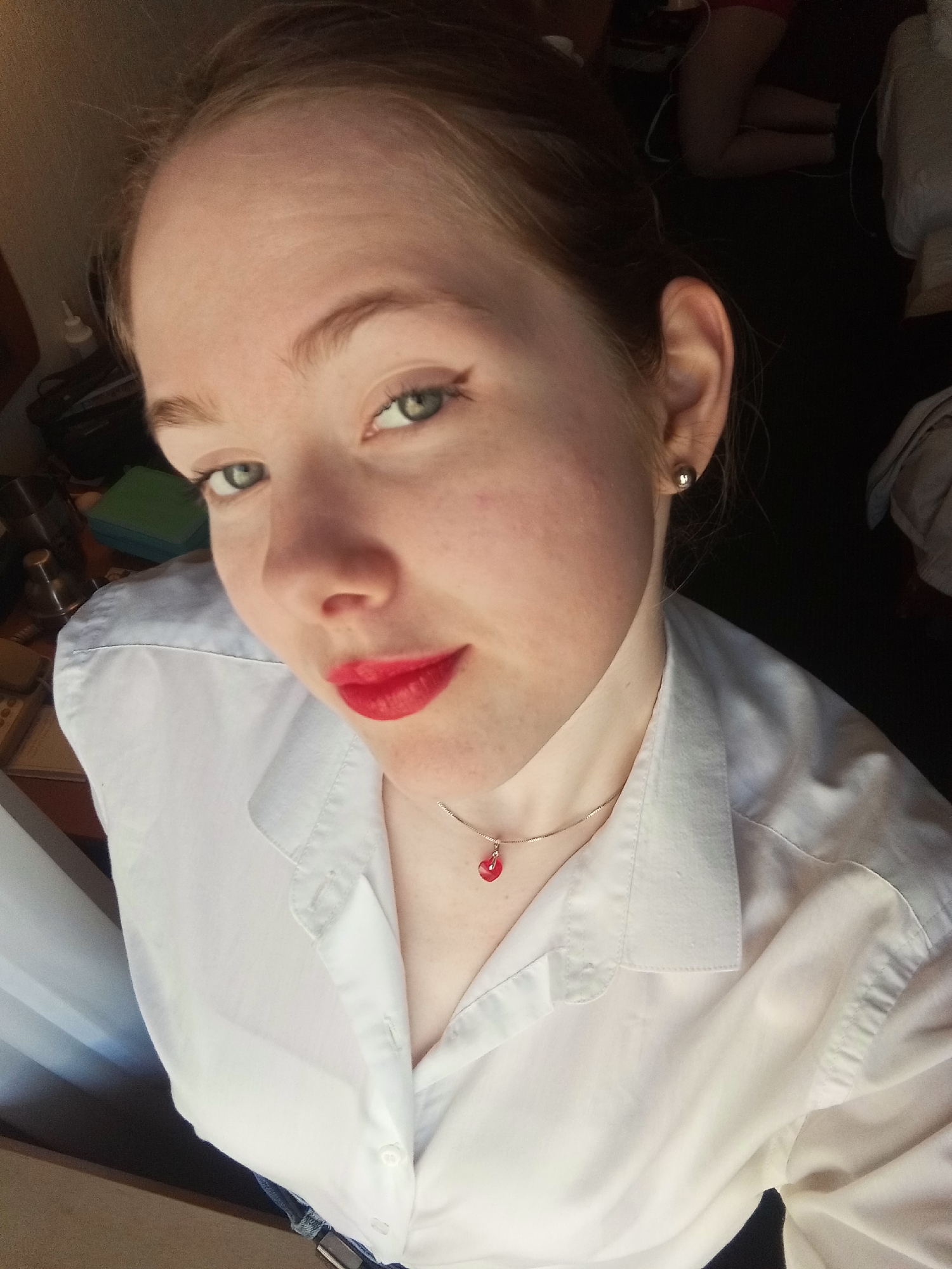 A young woman is positioned in a high-angle selfie, her face angled slightly to the left of the frame. She has fair skin, green eyes enhanced with subtle eyeliner, and red lipstick. Her hair is pulled back, revealing a silver necklace with a red pendant. She is wearing a white collared shirt.
In the background, there is a dark floor and indistinct objects, including what appears to be furniture, a window with a white curtain and an unidentified individual.
While it is not possible to determine the location as Saint Petersburg, Russia, from visual information alone, it is possible that additional information could be revealed via analysis of metadata, shadows, reflections or other contextual clues.