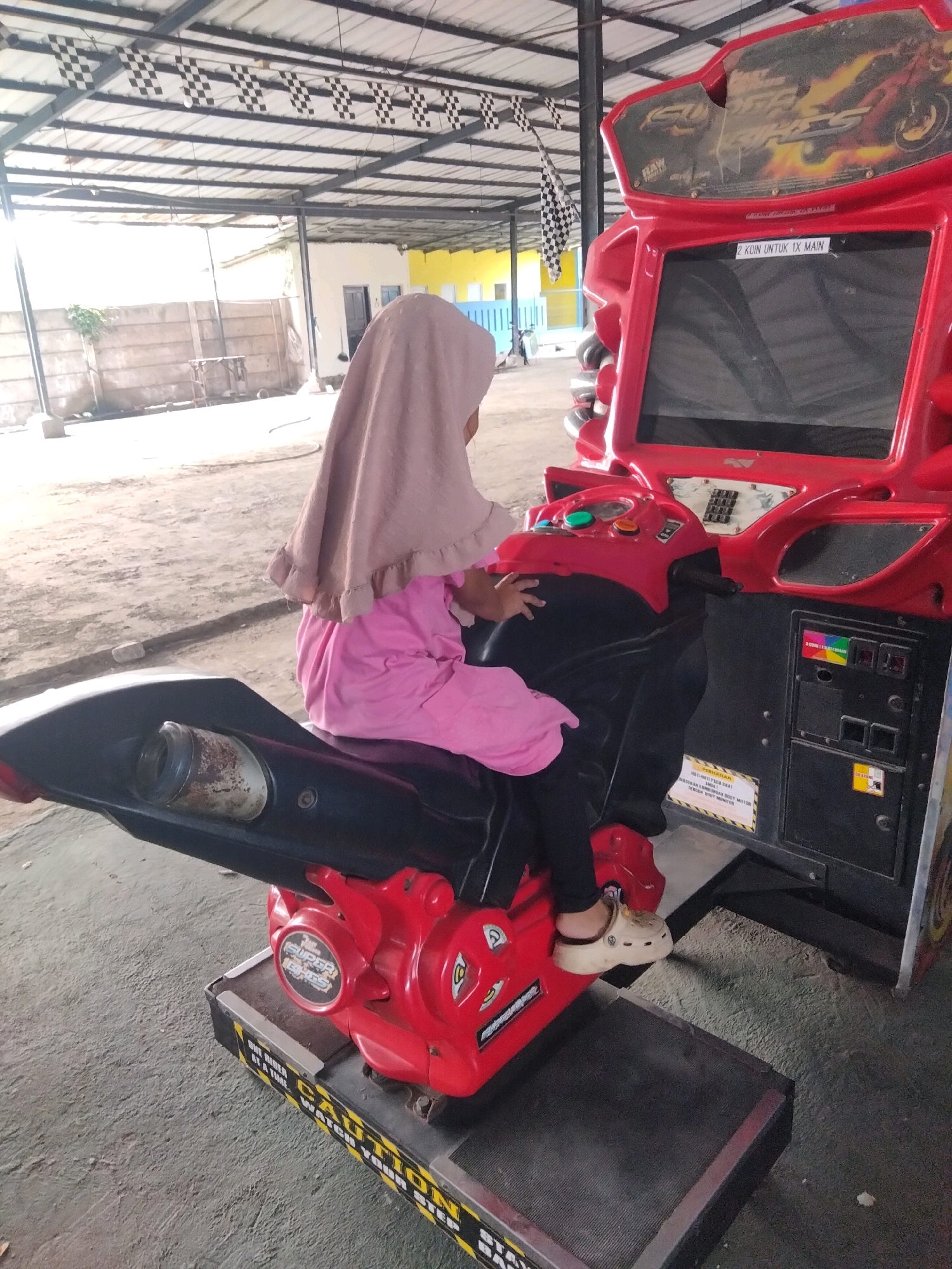 An Indonesian child wearing a pink hijab and a pink shirt is sitting on a red motorcycle arcade game. The child is wearing white Crocs. The arcade game is in an outdoor, covered area, possibly an amusement park or arcade. There are checkered flags hanging from the ceiling and on the arcade game. The ground is dirt and concrete. The arcade game has a screen that displays graphics of a motorcycle race. There is a sign on the arcade game that says "2 Koin Untuk 1X Main" which translates to "2 coins for 1 play." Another sign on the base of the arcade game says "CAUTION WATCH YOUR STEP." The environment appears to be a family-friendly entertainment venue in Sukaasih, Indonesia.