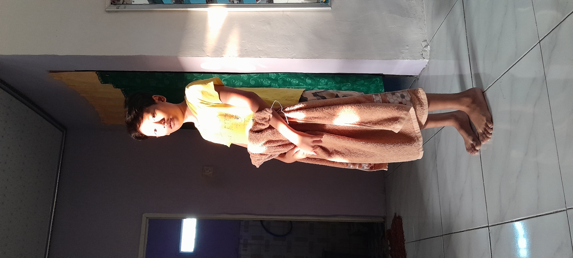 A young boy stands in what appears to be a room, possibly in Bendungan, Indonesia, bathed in the warm glow of sunlight streaming from the right. He is wearing a yellow t-shirt and patterned shorts, and is holding a folded brown towel. His eyes are open and he is looking slightly to his right, with his mouth slightly ajar. The room has tiled flooring and walls of muted colors, with a doorway visible on the left side leading to another area that is darker. The overall lighting suggests it is either sunrise or sunset.