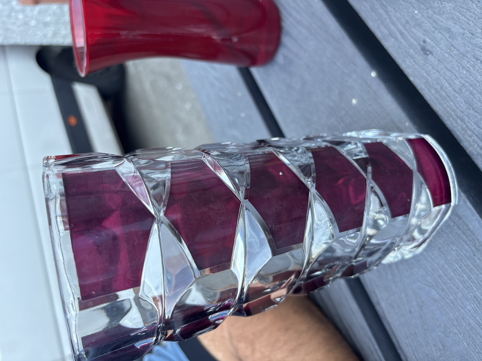 An outdoor, close-up, eye-level shot features two vases on a grey, slatted wooden table. The vase in the foreground is a tall, clear glass vase with a geometric, faceted design, revealing deep purple inserts within its structure. The vase on the right is a shorter, red glass vase with a curved silhouette. In the background, a white vehicle is partially visible. The scene appears to be set in the city of Boom, Belgium, specifically within the vicinity of a park or outdoor gathering area, suggested by the presence of similar tables and the overall ambiance.