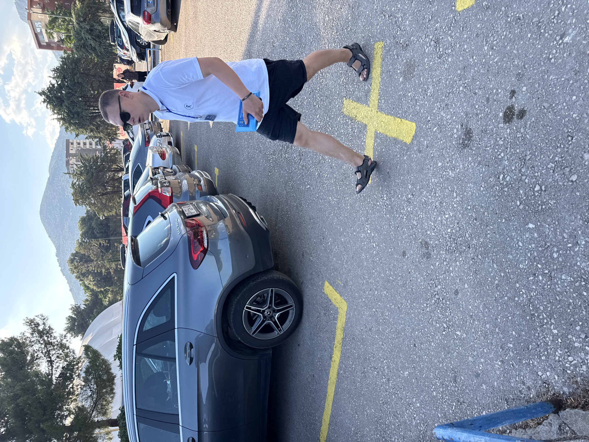 In Budva, Montenegro, a man walks through a parking lot. He is wearing a white polo shirt, black shorts, sunglasses, and dark sandals. He is holding a blue object in his right hand. The parking lot is paved with asphalt and marked with yellow lines. Several cars are parked in the lot, including a grey Mercedes-Benz SUV. In the background, there are trees, buildings, and mountains. The sky is blue with some clouds.