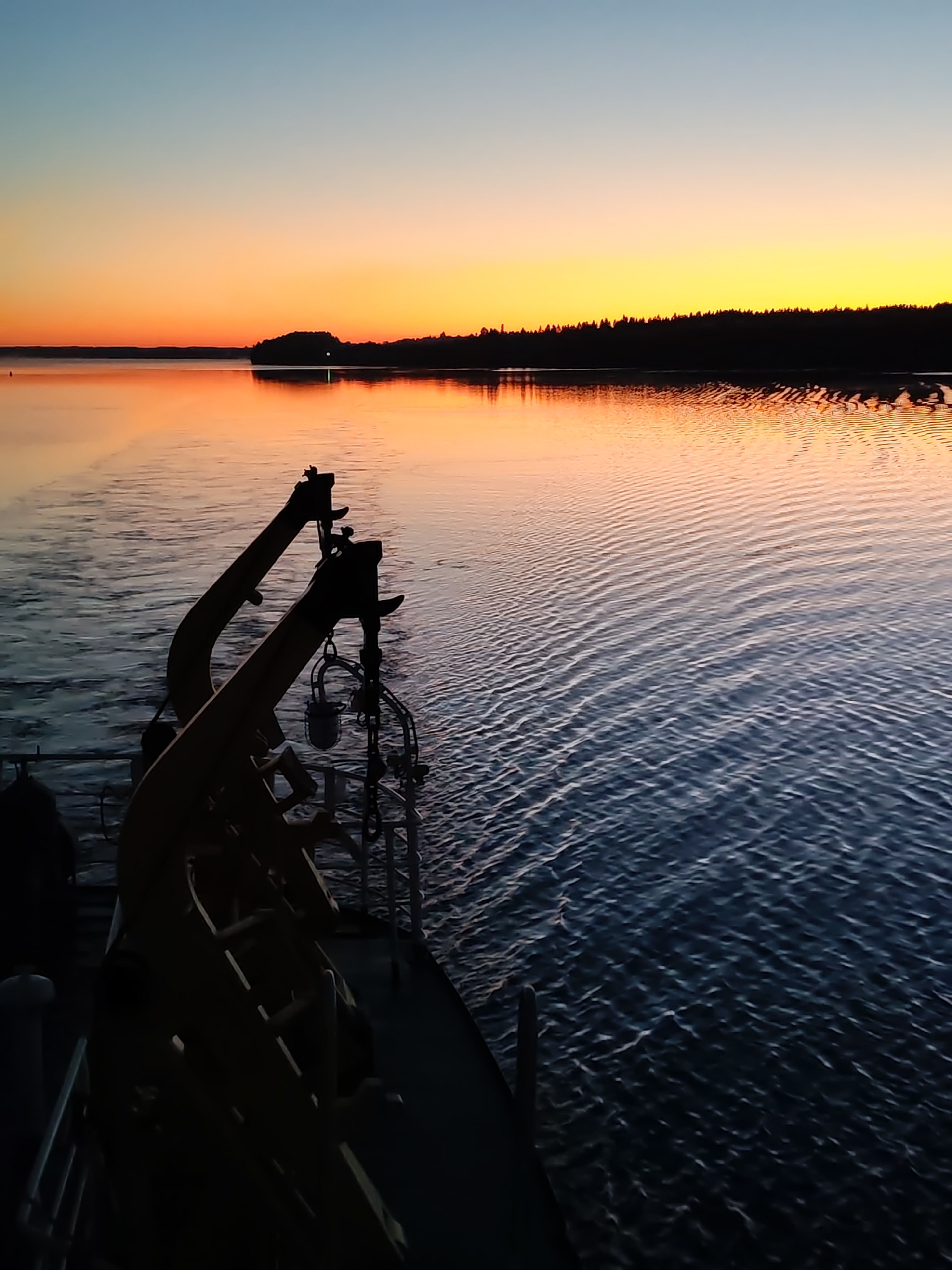 The image captures a sunset or sunrise over a large body of water in Khoroshevo, Russia. In the immediate foreground, the dark silhouette of a boat's deck is visible, featuring two prominent, curved, crane-like structures, possibly davits, on the starboard side. A small, lantern-like object hangs from one of these structures. No individuals are present. The water surface in the midground displays distinct ripples, reflecting the sky's colors, which transition from dark blue in the foreground to a vivid gradient of orange and pink closer to the horizon. In the background, a dark, densely forested landmass is silhouetted against the bright horizon, stretching horizontally across the scene. A faint point of light is discernible on this distant landmass. The sky above exhibits a clear gradient, moving from light blue at the top to intense orange and yellow directly above the horizon.
