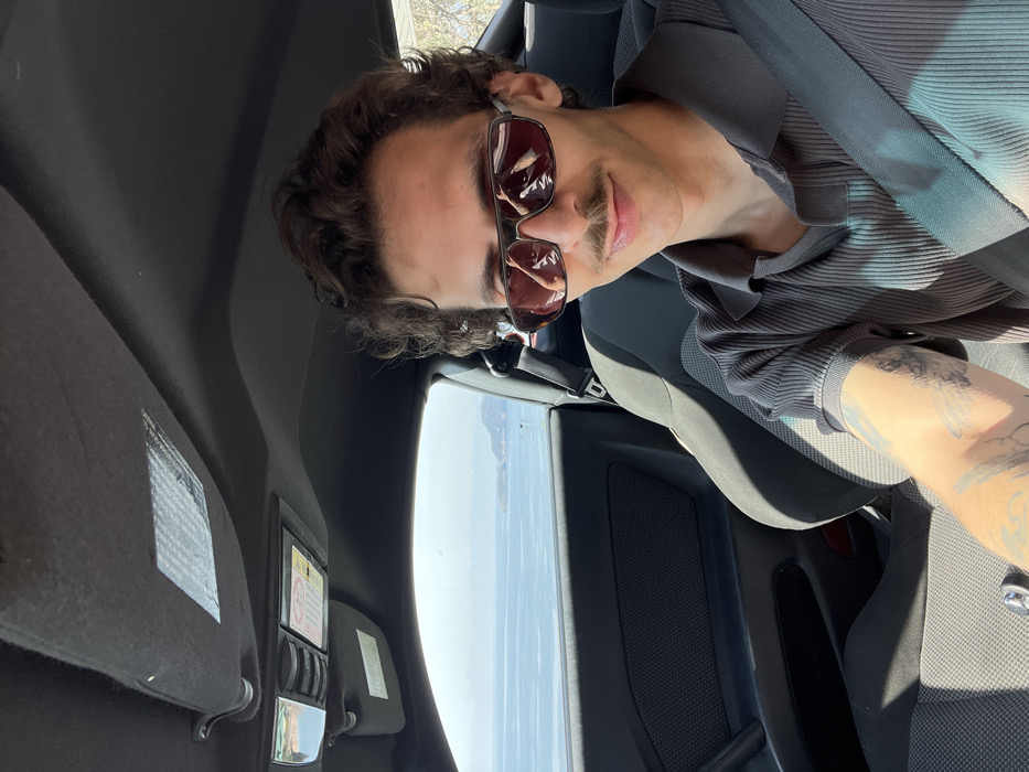 A male individual is depicted in a head-and-torso shot, seated within the dark interior of a vehicle, consistent with a driver's position. He has dark, curly hair and a prominent mustache, and wears dark-framed sunglasses with reflective reddish-brown lenses. He is dressed in a dark grey, short-sleeved, ribbed polo shirt, with a dark seatbelt secured across his chest. Tattoos are visible on his left forearm. The subject is looking directly at the camera with a slight smile. Through the open driver's side window, a wide expanse of calm, blue-grey sea is visible under a clear sky. Distant landmasses, characteristic of a coastal region, are present on the horizon, with one appearing as a small island or peninsula to the right. The scene is located in a coastal area, specifically consistent with Sveti Stefan, Montenegro. Vehicle interior details visible include a black headliner, a black sun visor, and an overhead console.