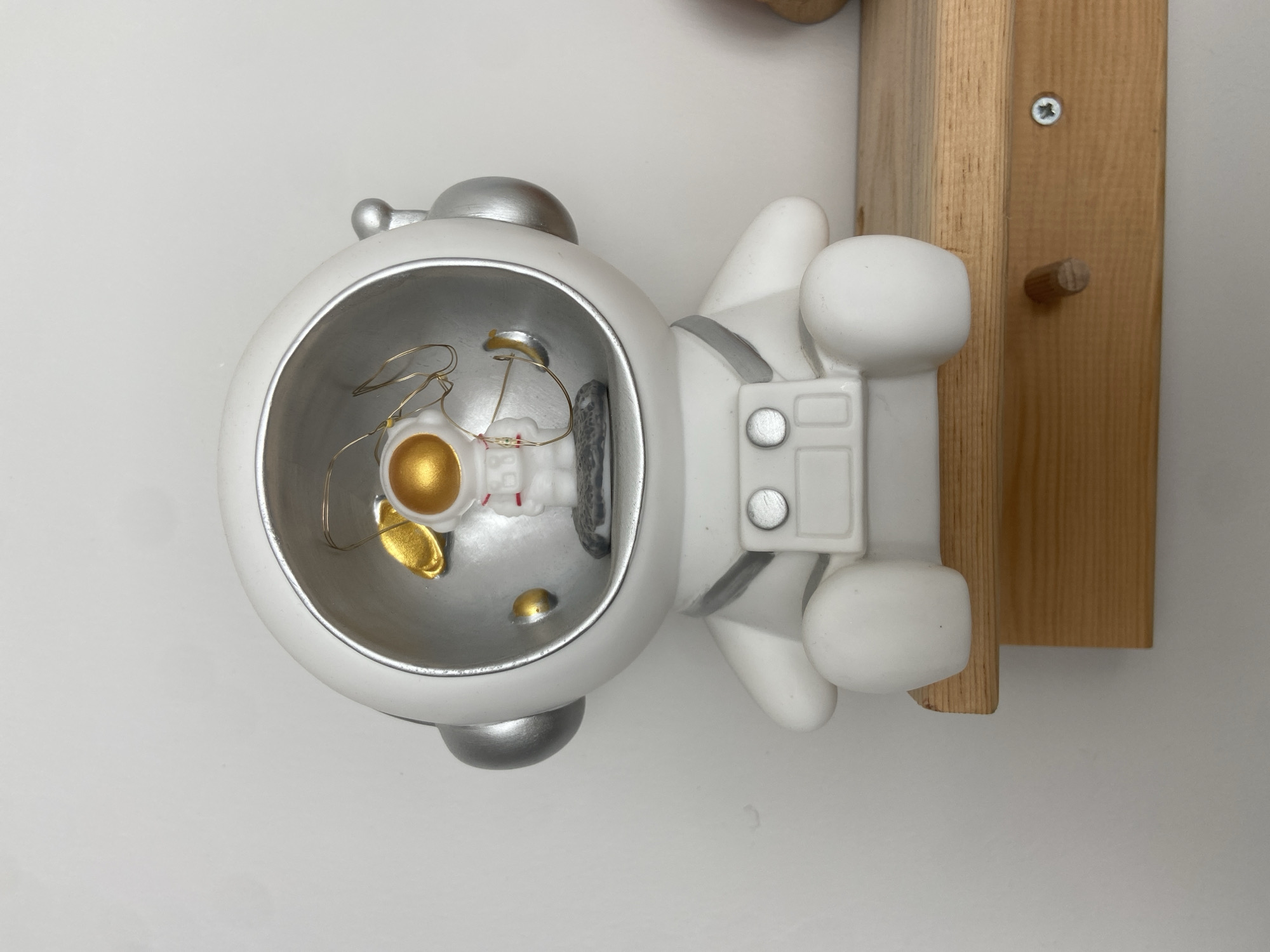 A white, seated astronaut figurine with a clear helmet containing small golden accents and thin wire lights is displayed on a light-colored wooden shelf against a plain white wall. The figurine has silver accents on its helmet and a detailed chest panel with two silver buttons. The shelf is supported by a piece of wood attached underneath. The setting appears to be indoors, possibly a home or a shop, and there is no discernible information to place it in Afşin, Turkey.