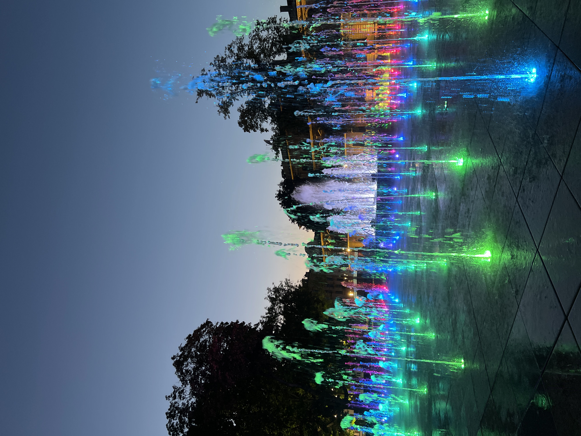 The image shows a colorful fountain display in Lublin, Poland, at dusk. Numerous jets of water are illuminated with vibrant blue, green, and pink lights, creating a dynamic and visually striking scene. The fountains are located in a paved square, likely a public space, with reflections of the lights shimmering on the wet surface. In the background, silhouetted trees frame the fountain area, and a building with illuminated windows can be seen behind the water jets. The sky is a deep blue, indicating twilight. There are no people visible in the image.